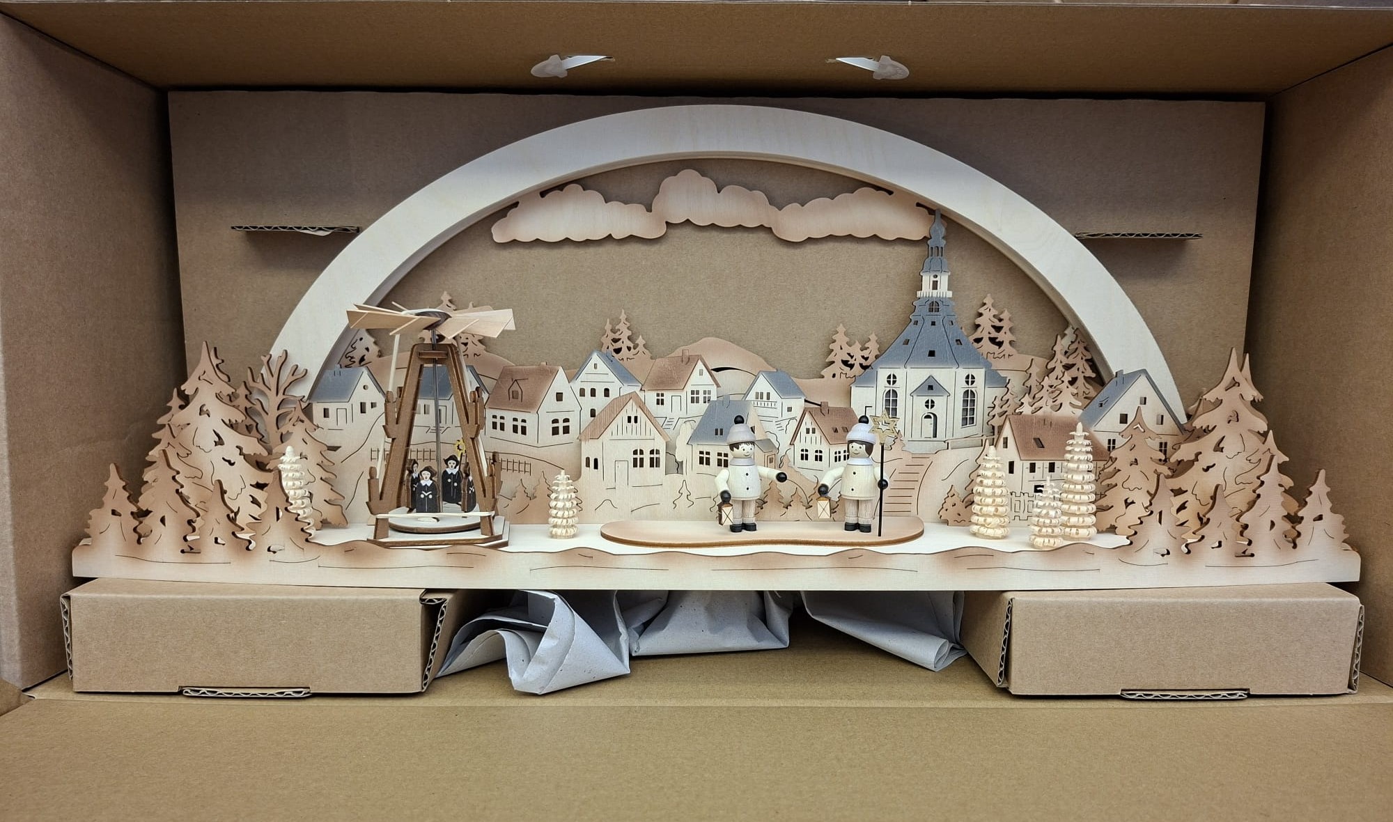 Ein traditioneller deutscher Weihnachtsbogen aus Holz mit einer detaillierten Dorfszene mit einer Kirche, Kiefern und kleinen Figuren, präsentiert in einer Aufbewahrungsbox aus Pappe.