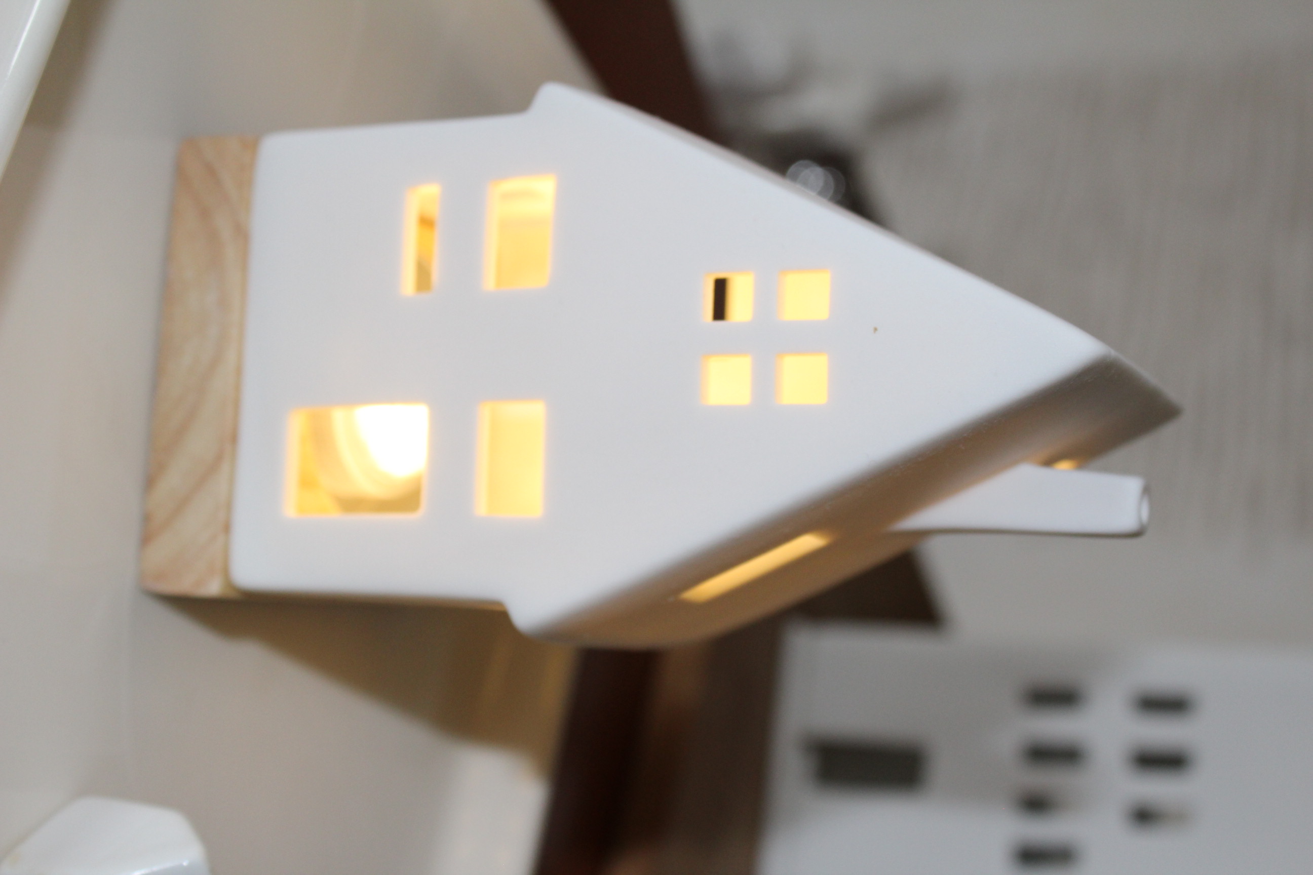 Ein bezaubernder hausförmiger Kerzenhalter aus weißer Keramik mit warmem gelbem Licht, das aus seinen Fenstern strömt und auf einem hellen Holzsockel ruht, perfekt für die Inneneinrichtung und Umgebungsbeleuchtung.