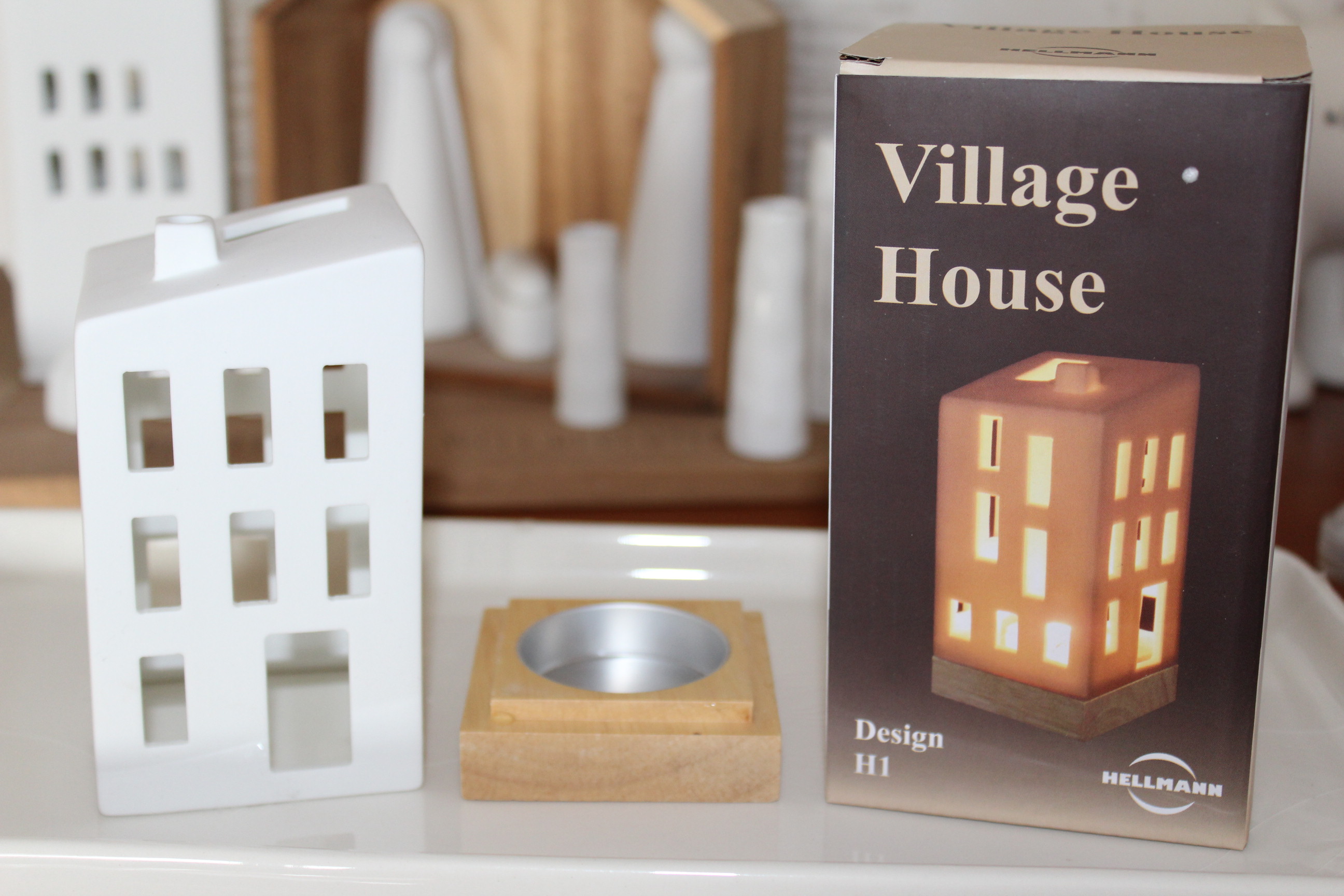 Eine weiße Dorfhausdekoration aus Keramik neben der Verpackung, die eine beleuchtete Version des Hauses zeigt. Vor der Schachtel steht ein Kerzenhalter aus Holz, im Hintergrund verschwimmen weitere weiße Keramikdekorationen.