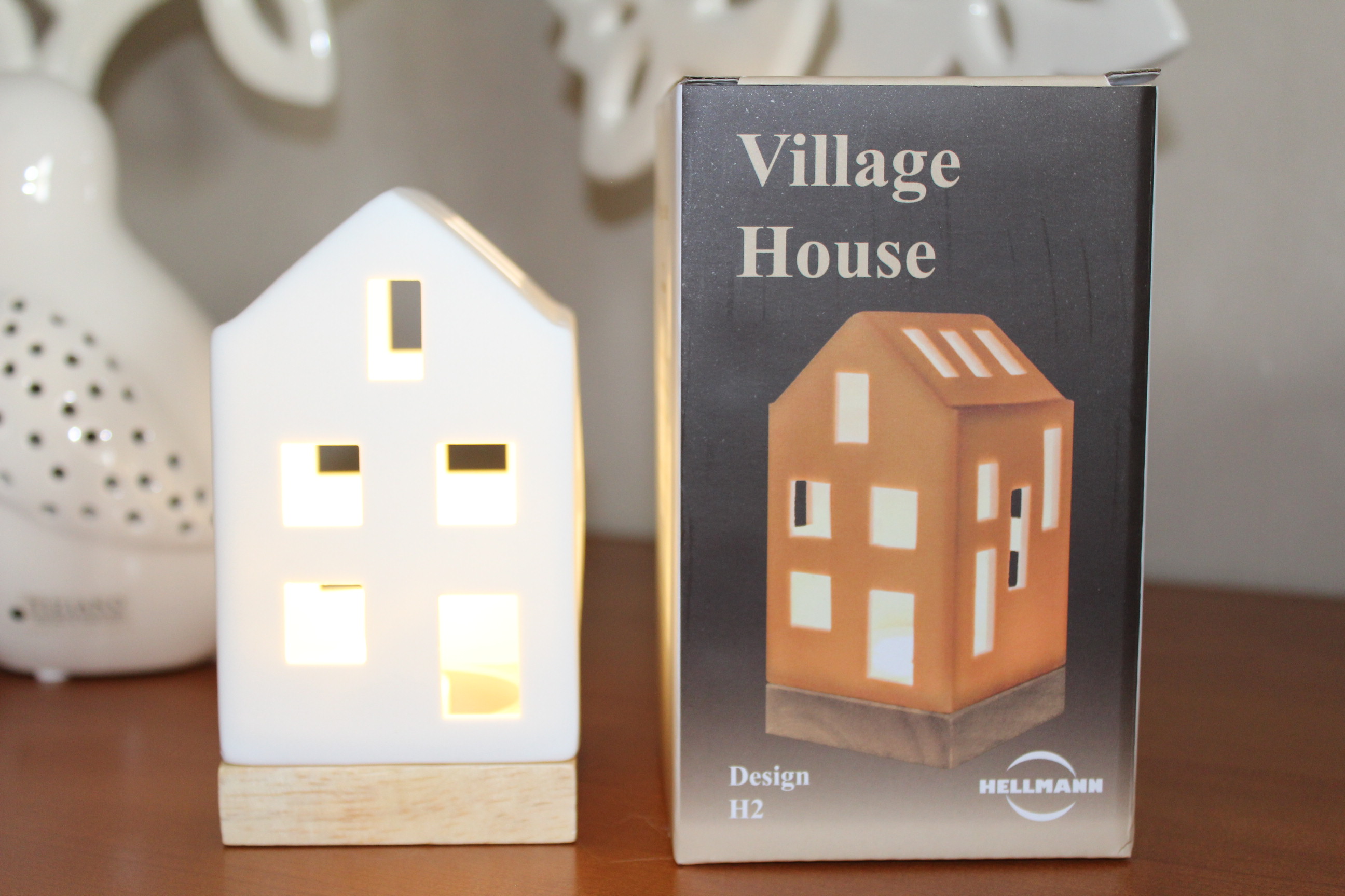 Ein weißer Dorfhaus-Kerzenhalter aus Keramik mit beleuchteten Fenstern steht neben seiner Verpackung mit der Aufschrift „Village House“ von Hellmann auf einer Holzoberfläche.