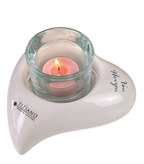 Herzförmiger, weiß glasierter Kerzenhalter mit einem Einsatz aus klarem Glas, in dem ein brennendes Teelicht steht. Auf der Oberfläche sind zwei Schriftzüge in schwarzer Schrift zu sehen: Einer mit dem Markennamen und einer mit einer Widmung.