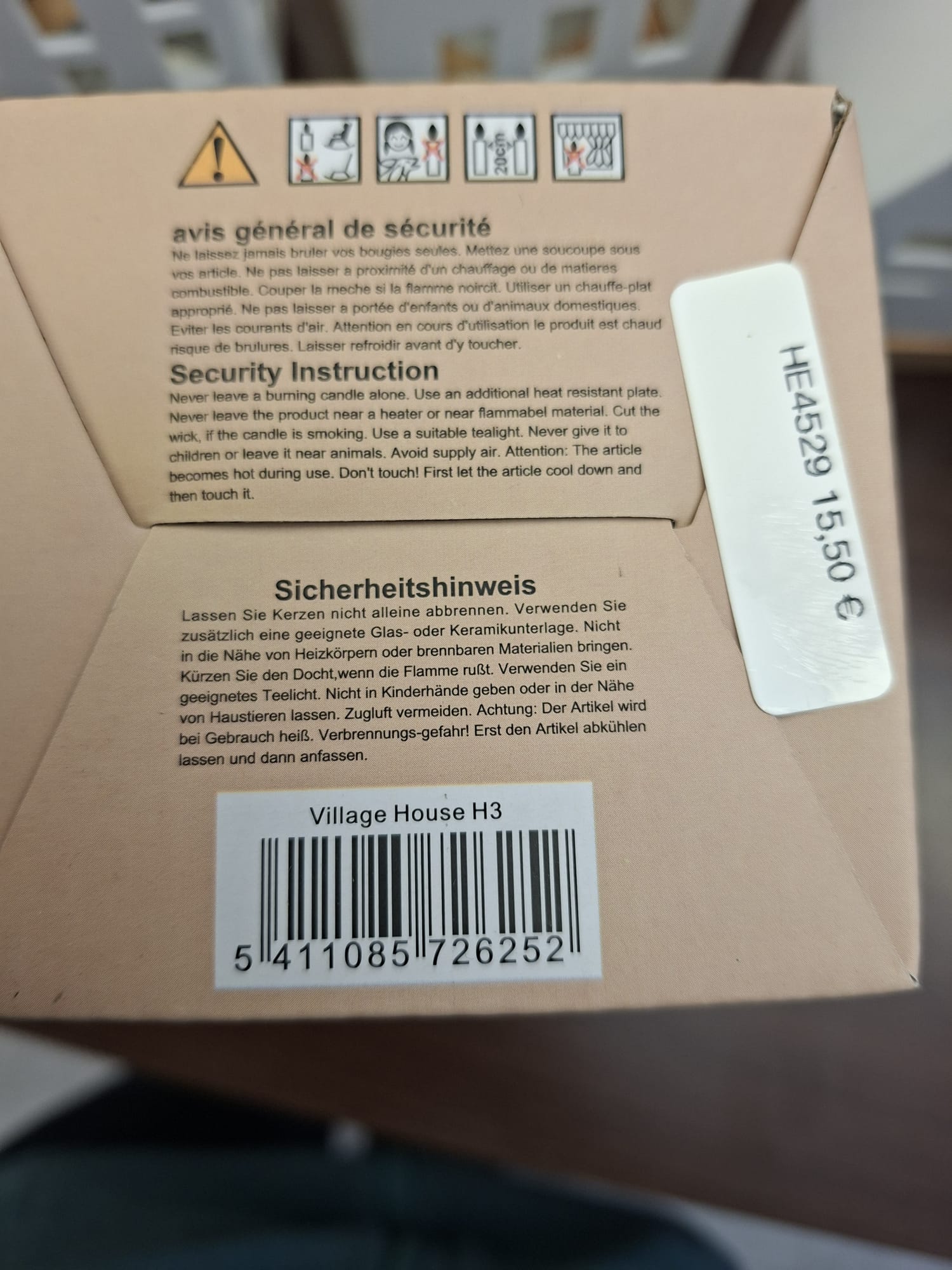 Nahaufnahme eines Kerzenwarnschilds mit Sicherheitshinweisen in Englisch, Französisch und Deutsch, mit universellen Sicherheitssymbolen und einem Barcode für die Kerze „Village House H3“.