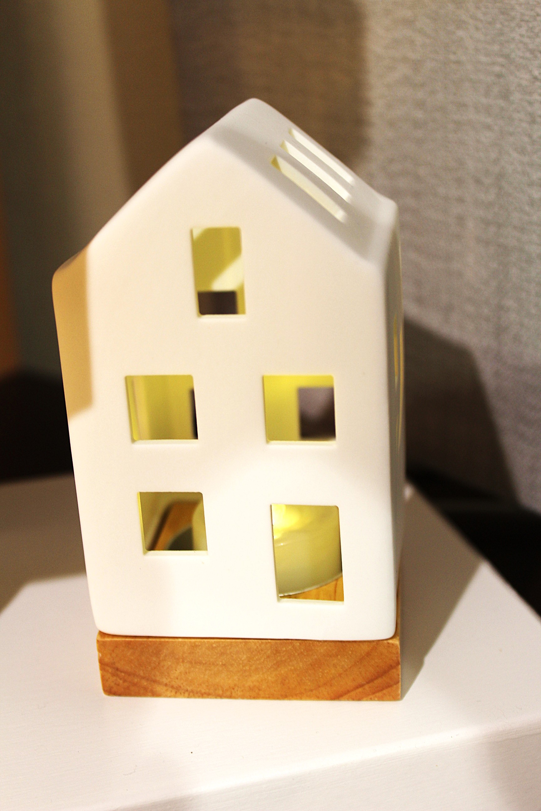 Weißer hausförmiger Kerzenhalter aus Keramik mit Holzsockel, sanft von innen beleuchtet vor einem neutralen Hintergrund.