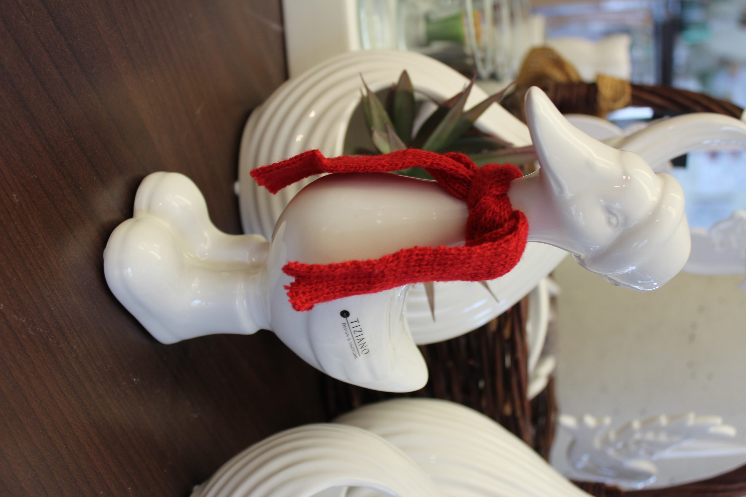 Eine weiße Keramik-Entenfigur mit passender weißer Mütze und leuchtend rotem Strickschal steht auf einer dunklen Holzoberfläche mit verschwommenen Wohnaccessoires im Hintergrund.
