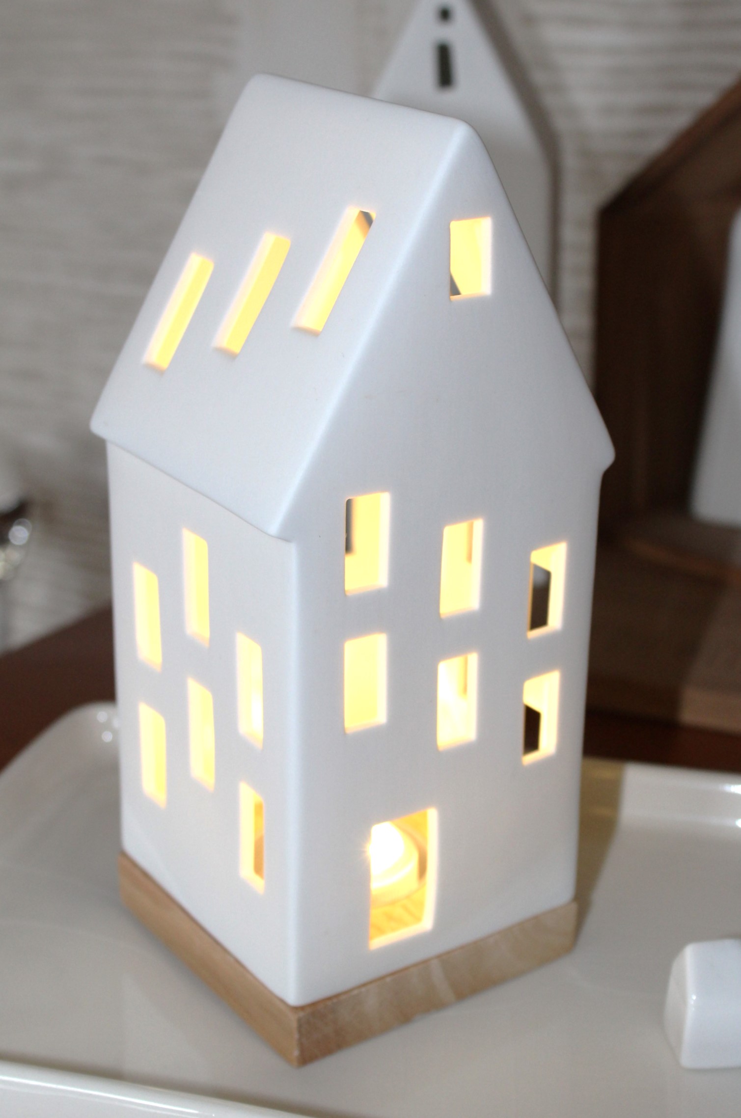 Ein weißer, hausförmiger Kerzenhalter aus Keramik mit beleuchteten Fenstern, der auf einem hellen Holzsockel ruht und eine warme und einladende Atmosphäre schafft.