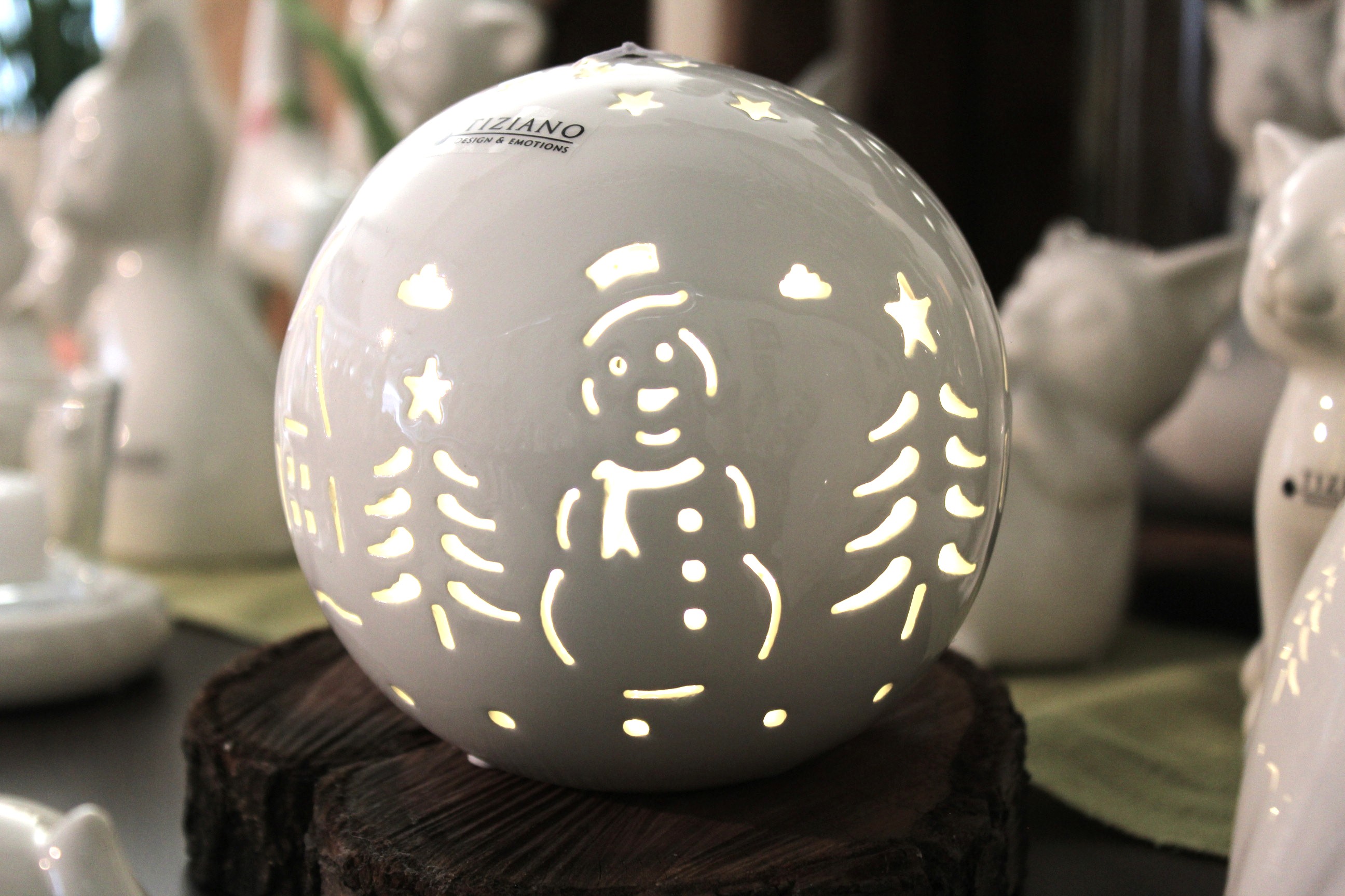 Eine weiße Rivello-Kugel aus Keramik von Tiziano Design & Emotions mit ausgeschnittenen Silhouetten eines Schneemanns, Weihnachtsbäumen und Sternen, die von innen durch ein LED-Licht beleuchtet wird und auf einem rustikalen Holzsockel steht.