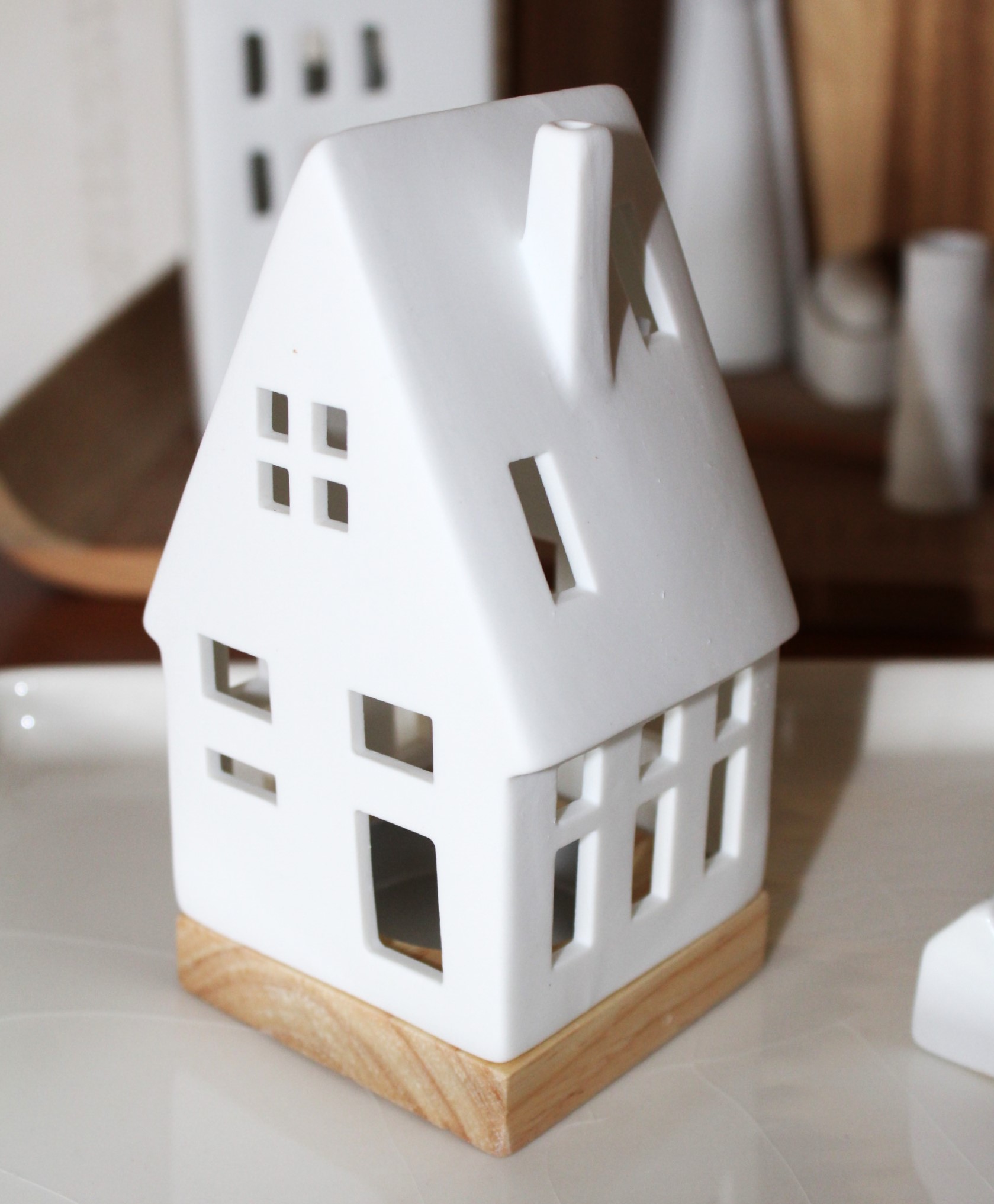 Eleganter Teelichthalter aus weißer Keramik mit Naturholzsockel, ausgeschnittenen Fenstern und einem Kamin, perfekt für die Inneneinrichtung und Umgebungsbeleuchtung.