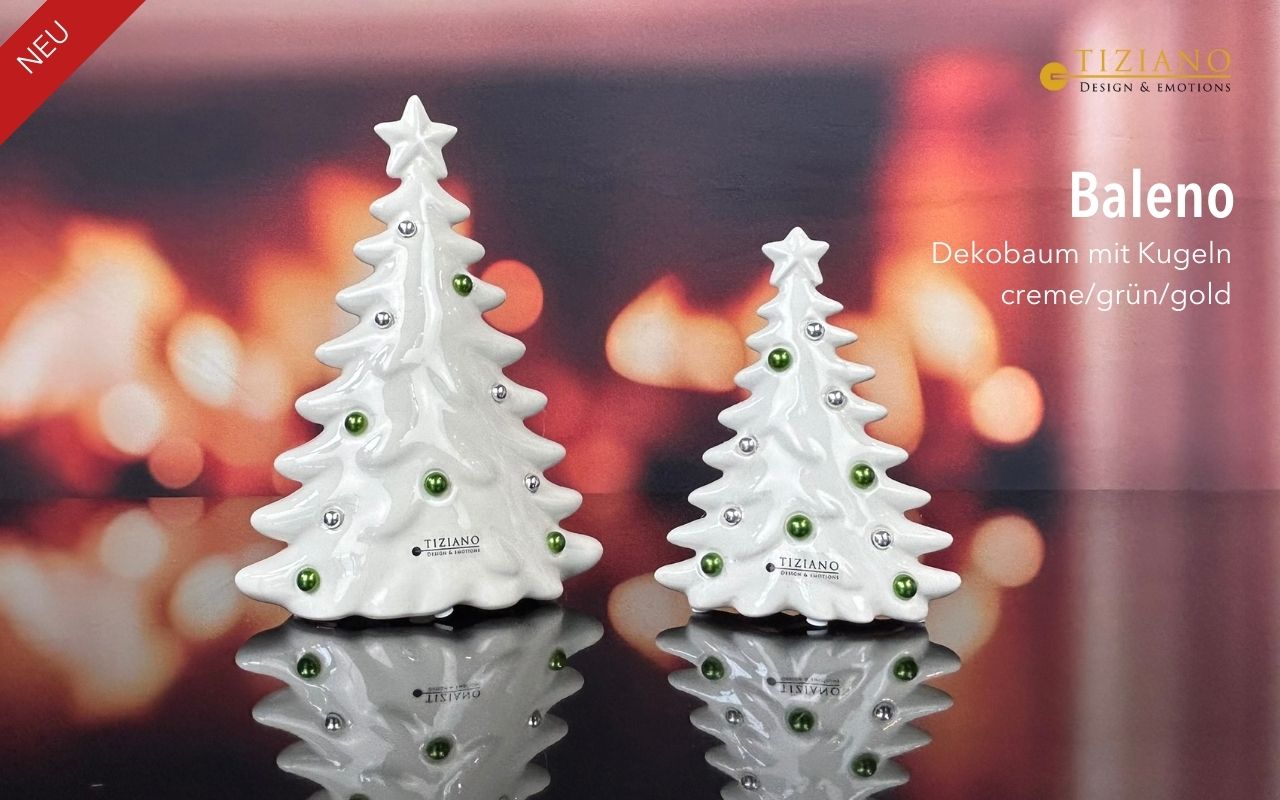 Zwei weiße Keramik-Weihnachtsbaumfiguren mit grünen und silbernen Zierkugeln, ausgestellt vor einem verschwommenen, warmen Kaminhintergrund mit Reflexionen auf einer dunklen Oberfläche.