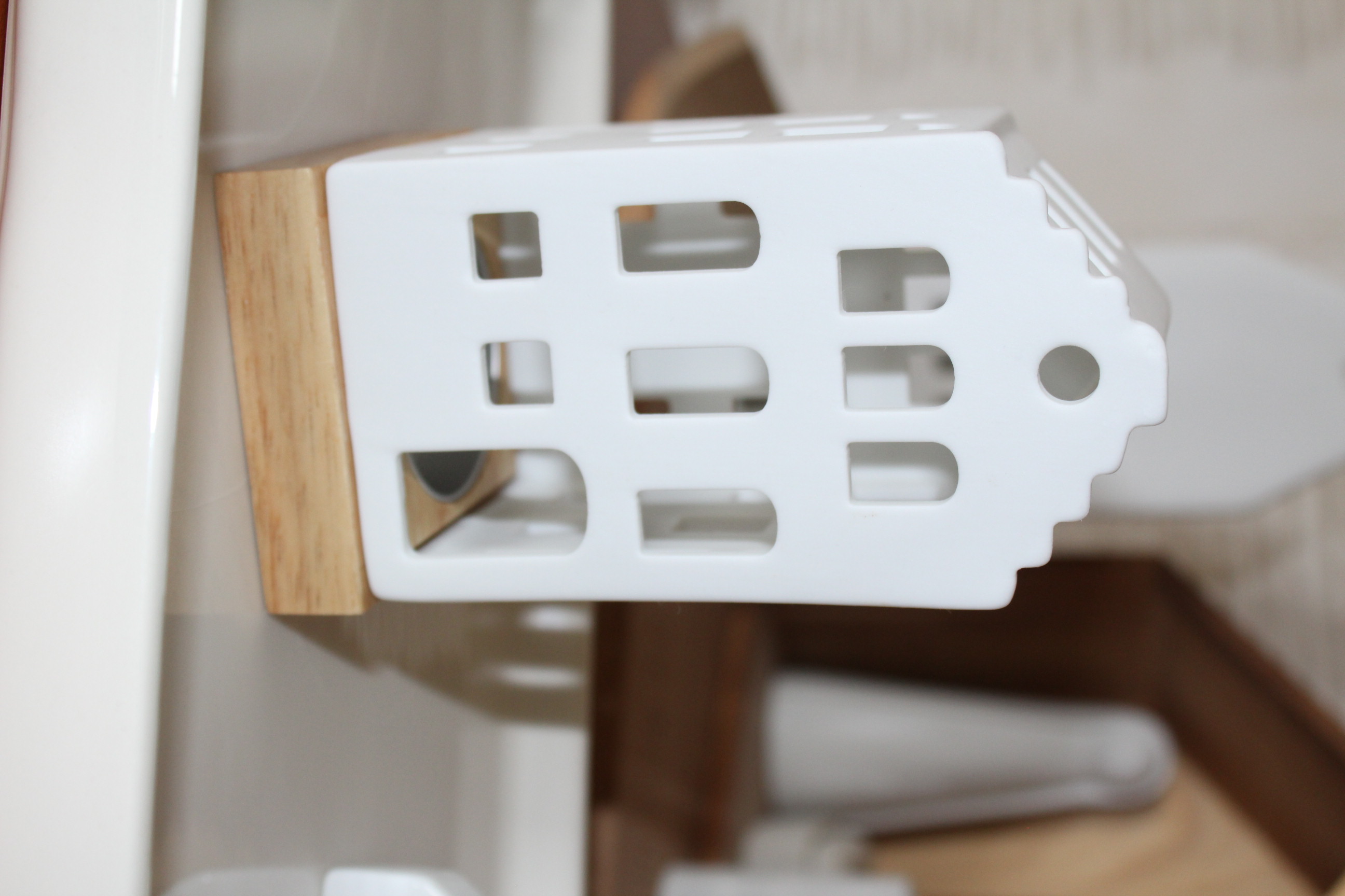 Eleganter hausförmiger Kerzenhalter aus weißer Keramik mit mehreren Fensterausschnitten, der auf einem hellen Holzsockel ruht und sich perfekt für die Inneneinrichtung und Umgebungsbeleuchtung eignet.