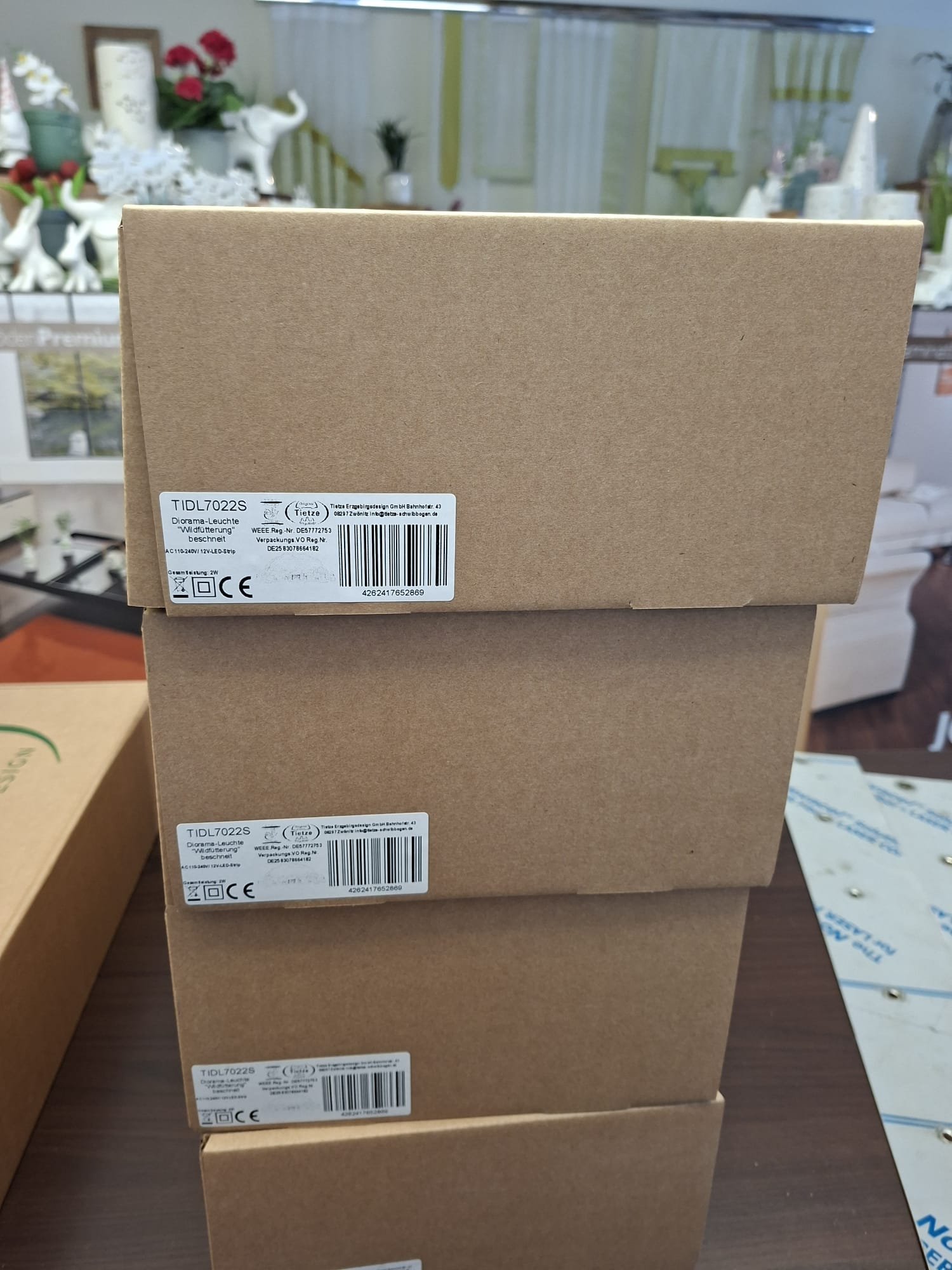 Ein Stapel aus vier braunen Kartons mit jeweils einem weißen Produktetikett mit Barcode, der Modellnummer TIDL7022S und Herstellerangaben vor dem verschwommenen Hintergrund der Inneneinrichtung eines Einzelhandelsgeschäfts.