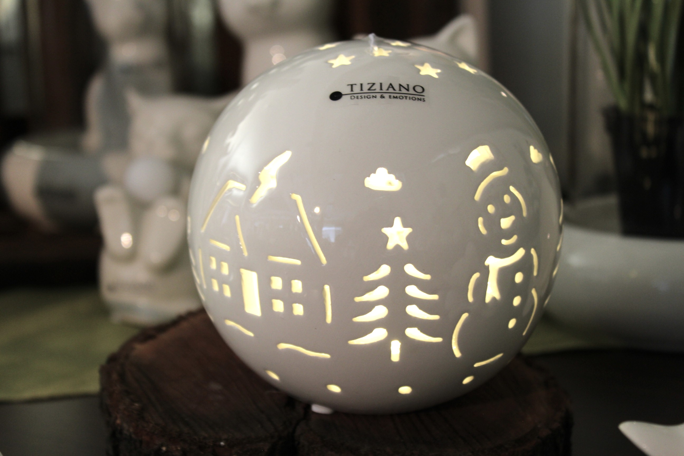 Eine weiße Keramikkugellampe von Tiziano Design & Emotions mit ausgeschnittenen Wintermotiven, darunter ein Schneemann, ein Weihnachtsbaum und ein Haus, die von innen durch ein LED-Licht beleuchtet wird und auf einem rustikalen Holzsockel ruht.