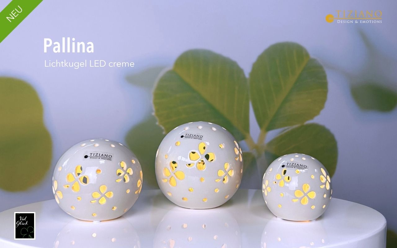 Drei cremefarbene LED-Lichtkugeln von Tiziano Pallina mit zarten Blumenausschnitten, die einen warmen Glanz ausstrahlen, angeordnet auf einer weißen Oberfläche mit verschwommenem grünem Blattwerk im Hintergrund, präsentieren eine elegante Inneneinric