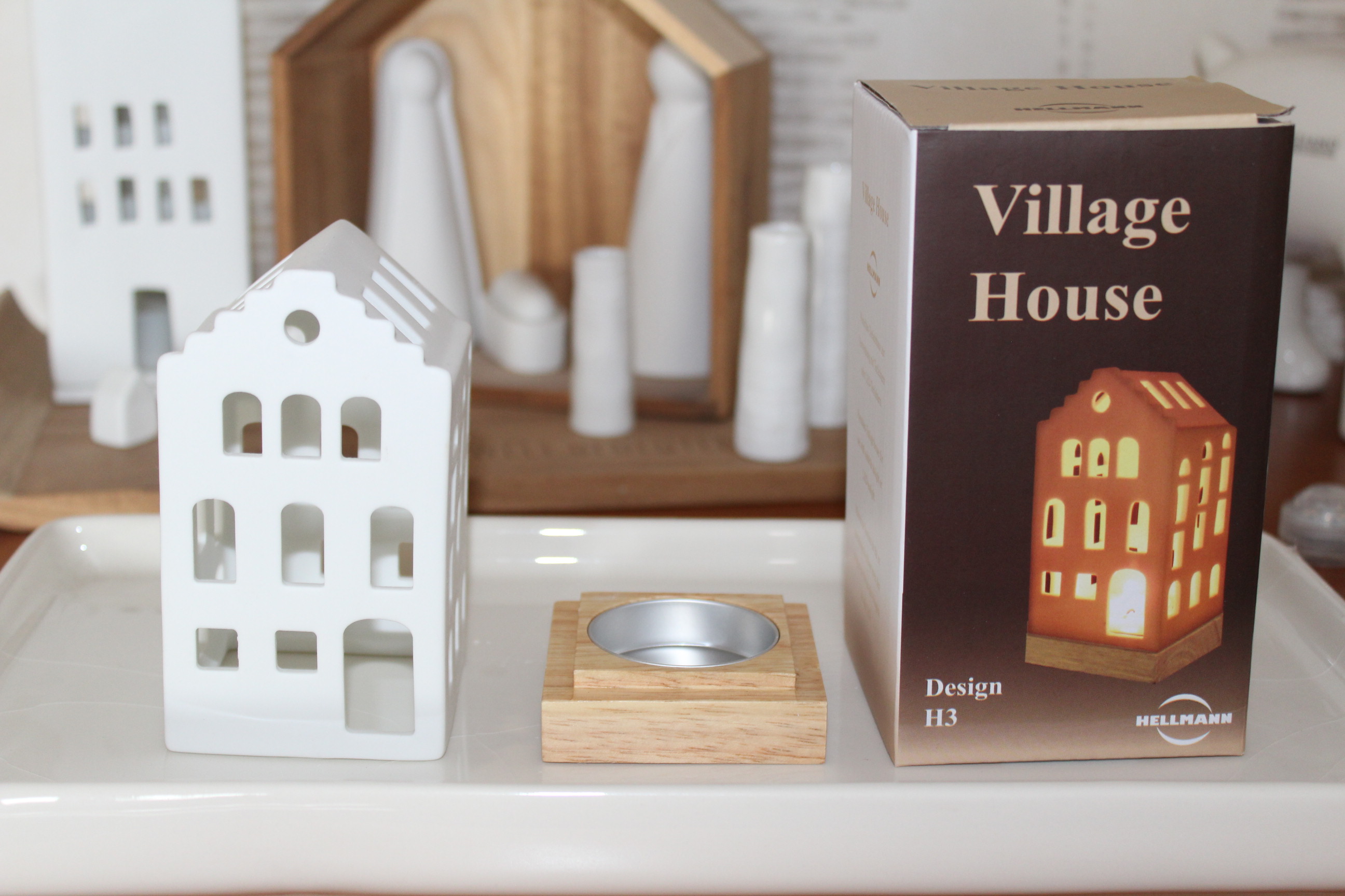 Ein weißer Dorfhaus-Kerzenhalter aus Keramik, ein Holzsockel für ein Teelicht und eine „Village House“-Produktbox von Hellmann. Ideal für Heimdekoration und festliche Präsentationen.