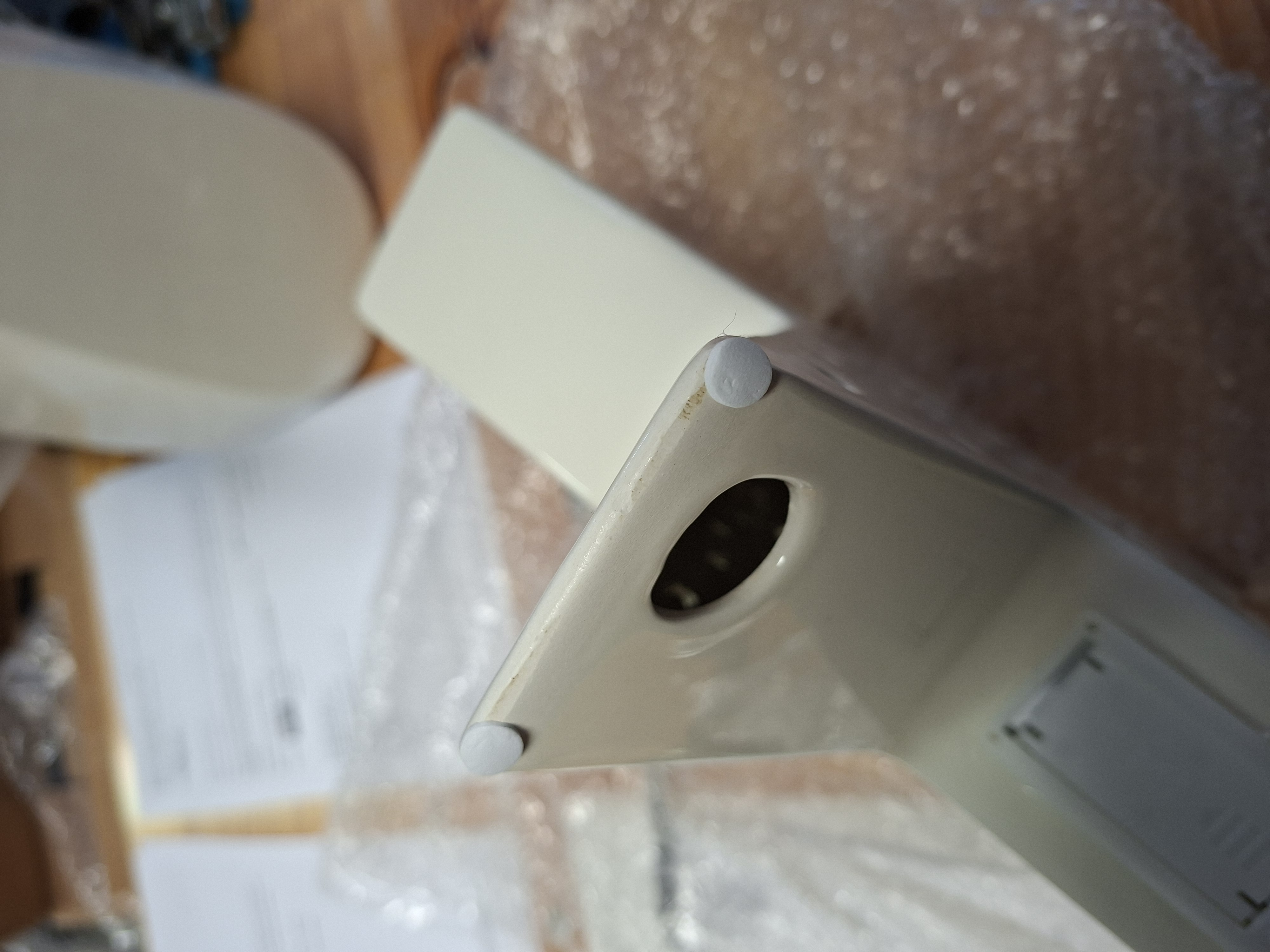 Nahaufnahme eines weißen elektronischen Geräts mit abgerundeter Kante und Anschluss. Der Hintergrund zeigt Dokumente und Luftpolsterfolie, die auf das kürzlich erfolgte Auspacken hinweisen.