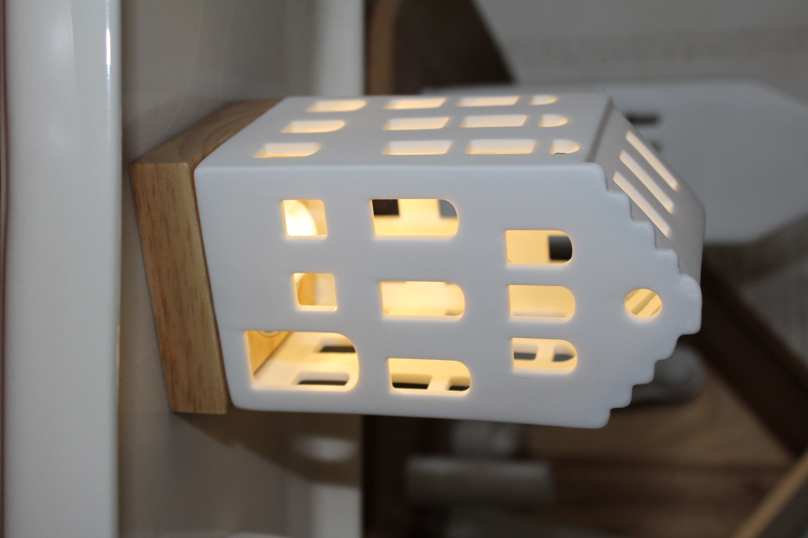 Eleganter hausförmiger Kerzenhalter aus weißer Keramik mit beleuchteten Fenstern, der auf einem Sockel aus Naturholz ruht und sich perfekt für die Inneneinrichtung und Umgebungsbeleuchtung eignet.