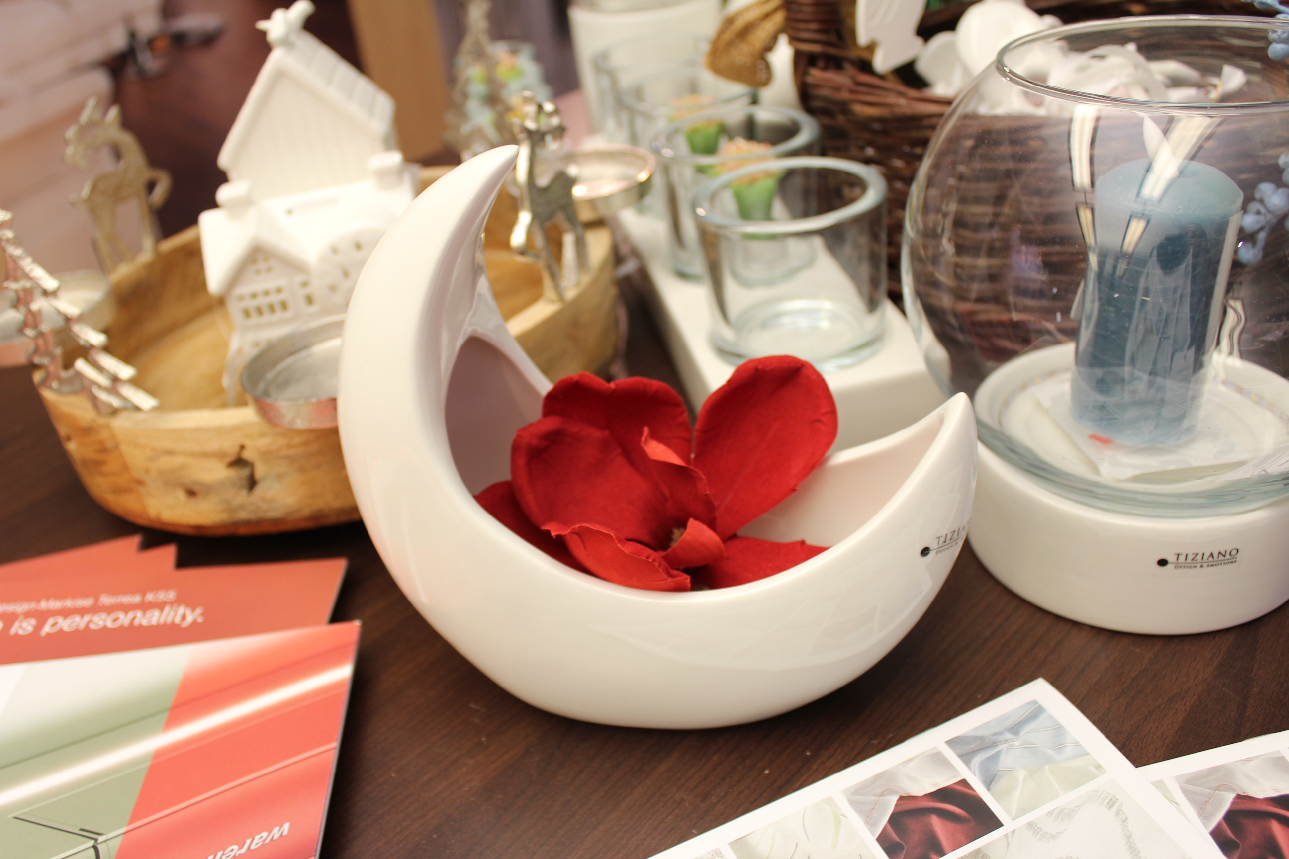 Ein cremefarbener Keramik-Übertopf von Tiziano Design & Emotions Marina in Form einer Mondsichel mit einer großen roten Blume, ausgestellt auf einer dunklen Holzoberfläche neben anderen saisonalen Wohnaccessoires wie Kerzen und einem Holztablett.