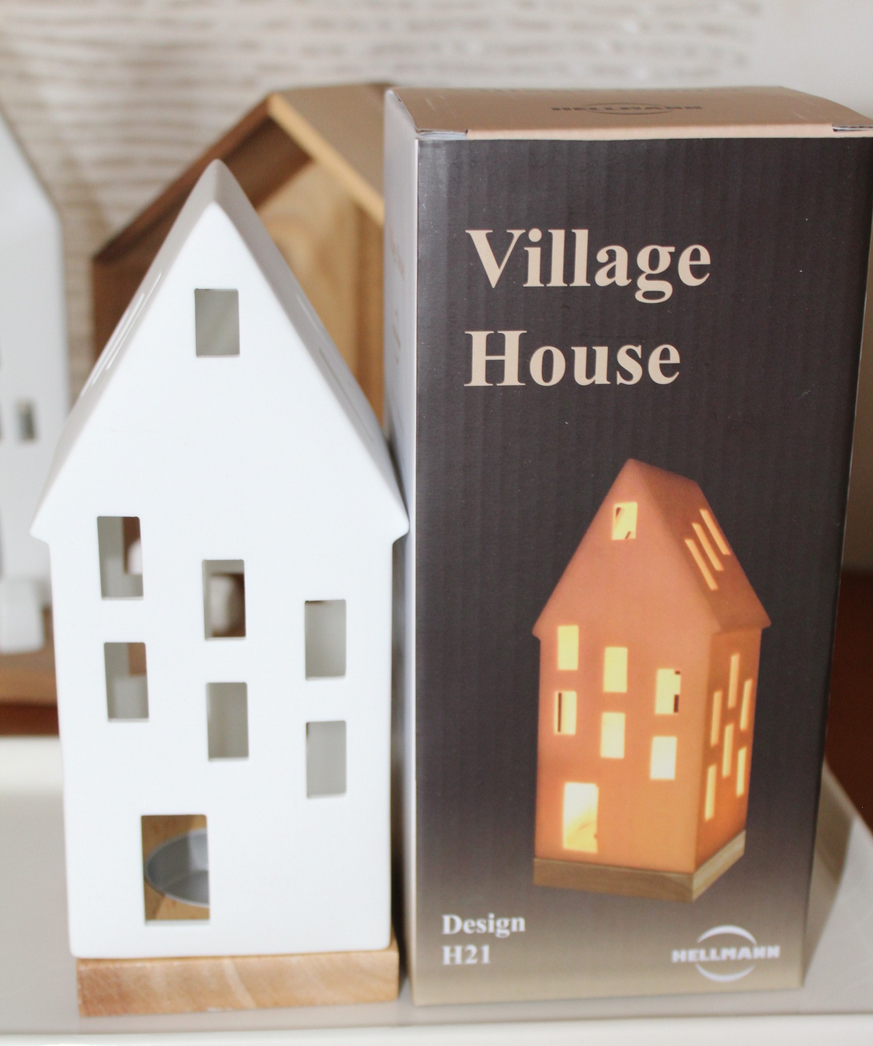 Neben der Verpackung steht ein weißer Dorfhaus-Kerzenhalter aus Keramik mit Holzsockel, auf dem ein beleuchtetes orangefarbenes Keramikhaus und der Text „Village House Design H21 HELLMANN“ zu sehen sind.