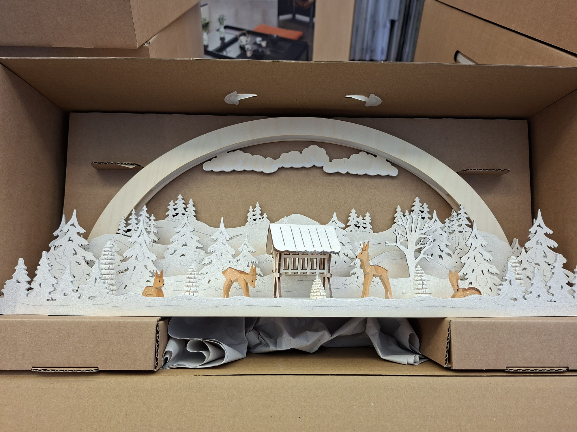 Eine Weihnachtsbogendekoration aus Holz mit einer Winterwaldszene mit Hirschen, einem Futtertrog und schneebedeckten Bäumen, präsentiert in einer Versandschachtel aus Pappe.