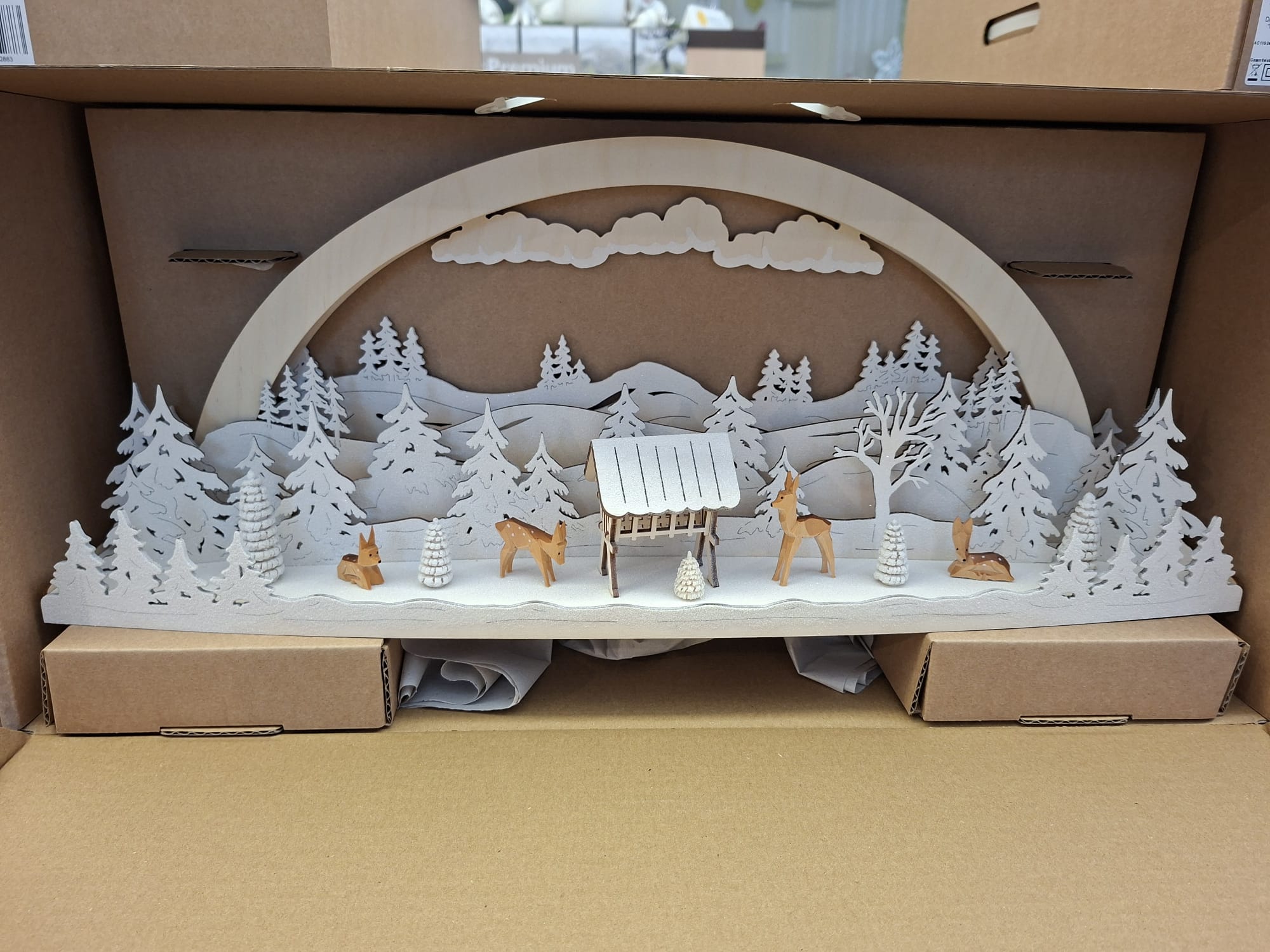 Ein dekorativer Weihnachtsbogen aus Holz mit einer Winterwaldszene mit weißen Bäumen, einem kleinen Futtertrog und mehreren kleinen Hirschfiguren aus Holz, alles verpackt in einer Pappschachtel.