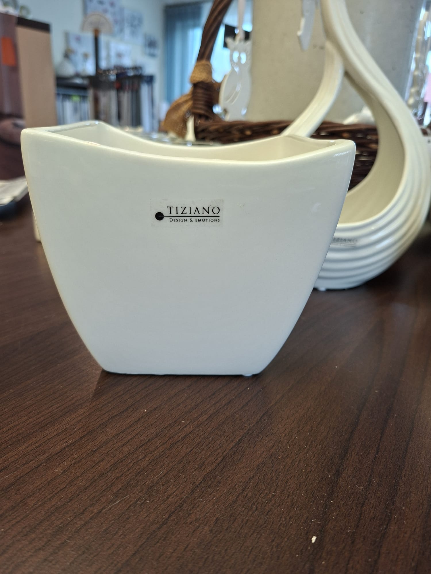 Ein Pflanzgefäß aus weißer Keramik der Marke Tiziano steht auf einem dunklen Holztisch, im verschwommenen Hintergrund sind eine dekorative schwanenförmige Vase aus weißer Keramik und ein Weidenkorb zu sehen.
