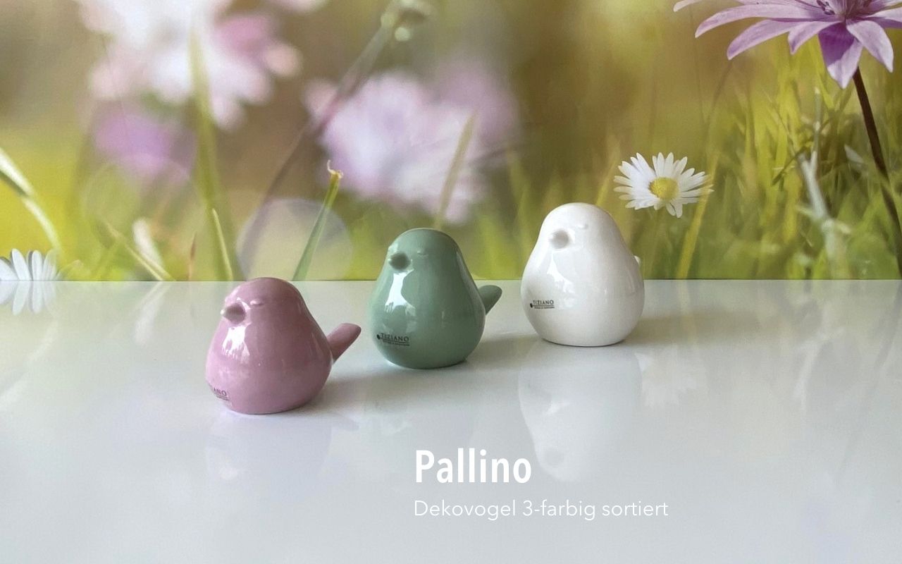 Drei kleine, glatte Vogelfiguren aus Keramik in Pastellrosa, Hellgrün und Weiß, angeordnet auf einer reflektierenden weißen Oberfläche vor einem verschwommenen Blumenhintergrund.