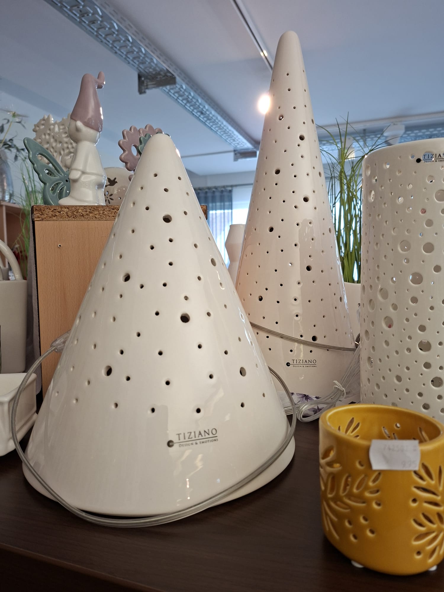 Zwei weiße, kegelförmige Tiziano-Keramiklampen mit perforierten Löchern stehen auf einem Regal neben einer zylindrischen weißen Lampe und einem kleinen gelben dekorativen Kerzenhalter, im Hintergrund ist eine kleine Zwergenfigur zu sehen.
