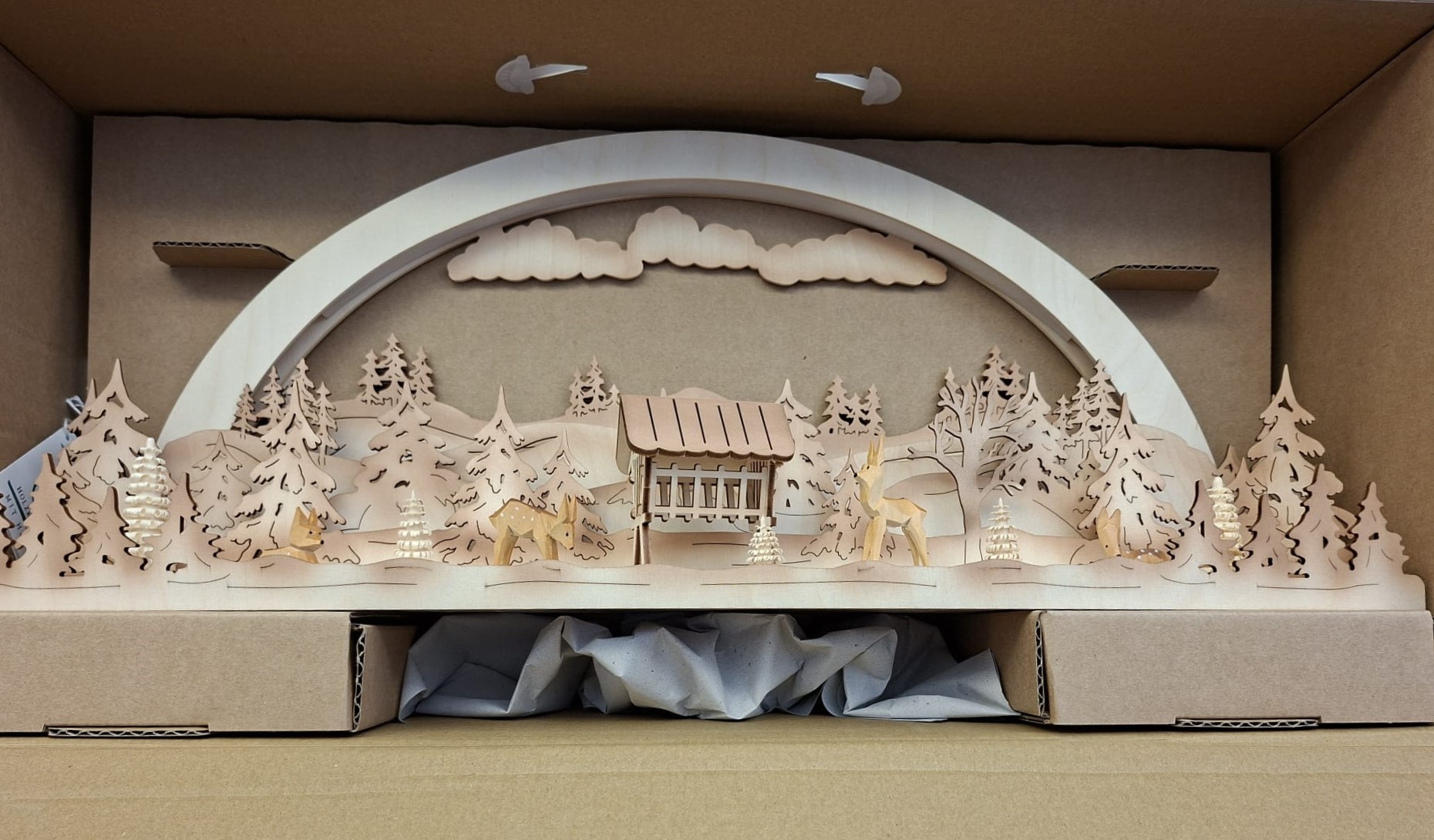 Ein dekorativer Schwibbogen aus Holz mit einer lasergeschnittenen Winterwaldszene mit Hirschen, einem Futtertrog und Kiefern, zum Schutz in einer Pappschachtel verpackt.