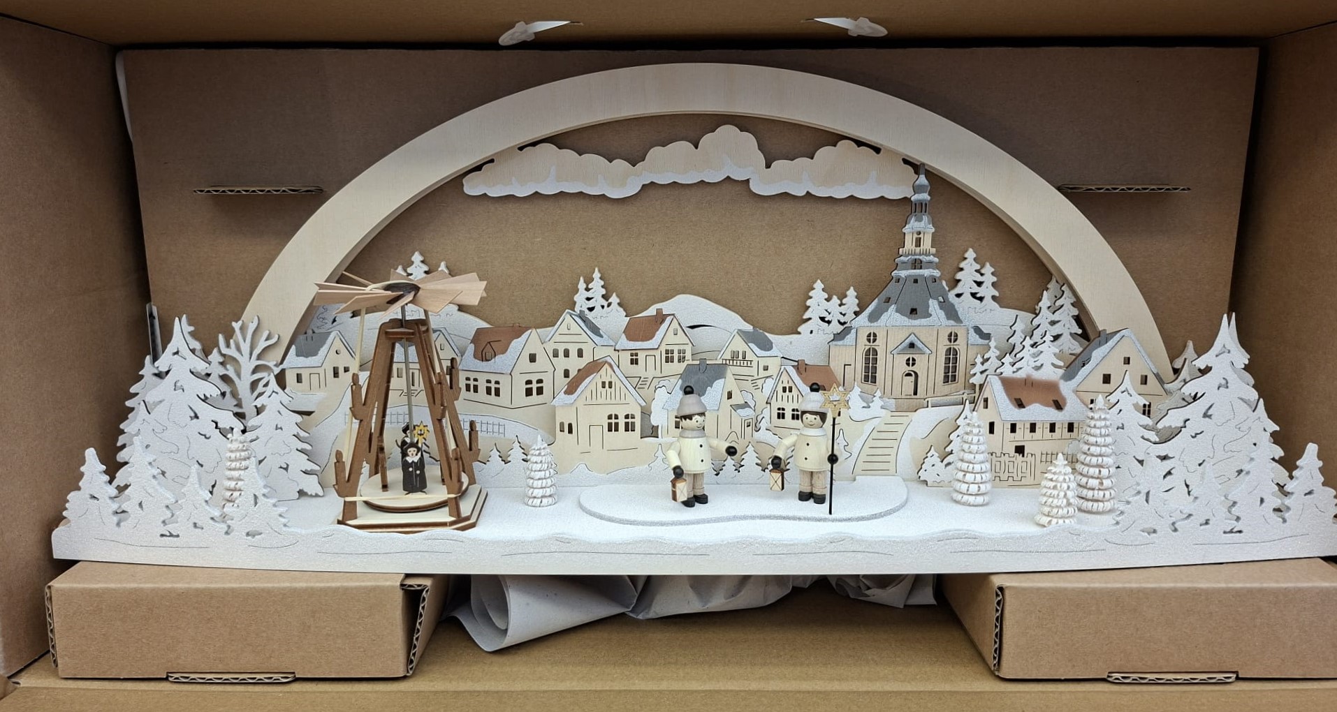 Ein traditioneller Weihnachtsbogen aus Holz mit einer detaillierten Winterdorfszene mit einer Kirche, schneebedeckten Bäumen und kleinen Figuren mit Laternen, präsentiert in einer Versandschachtel aus Pappe.