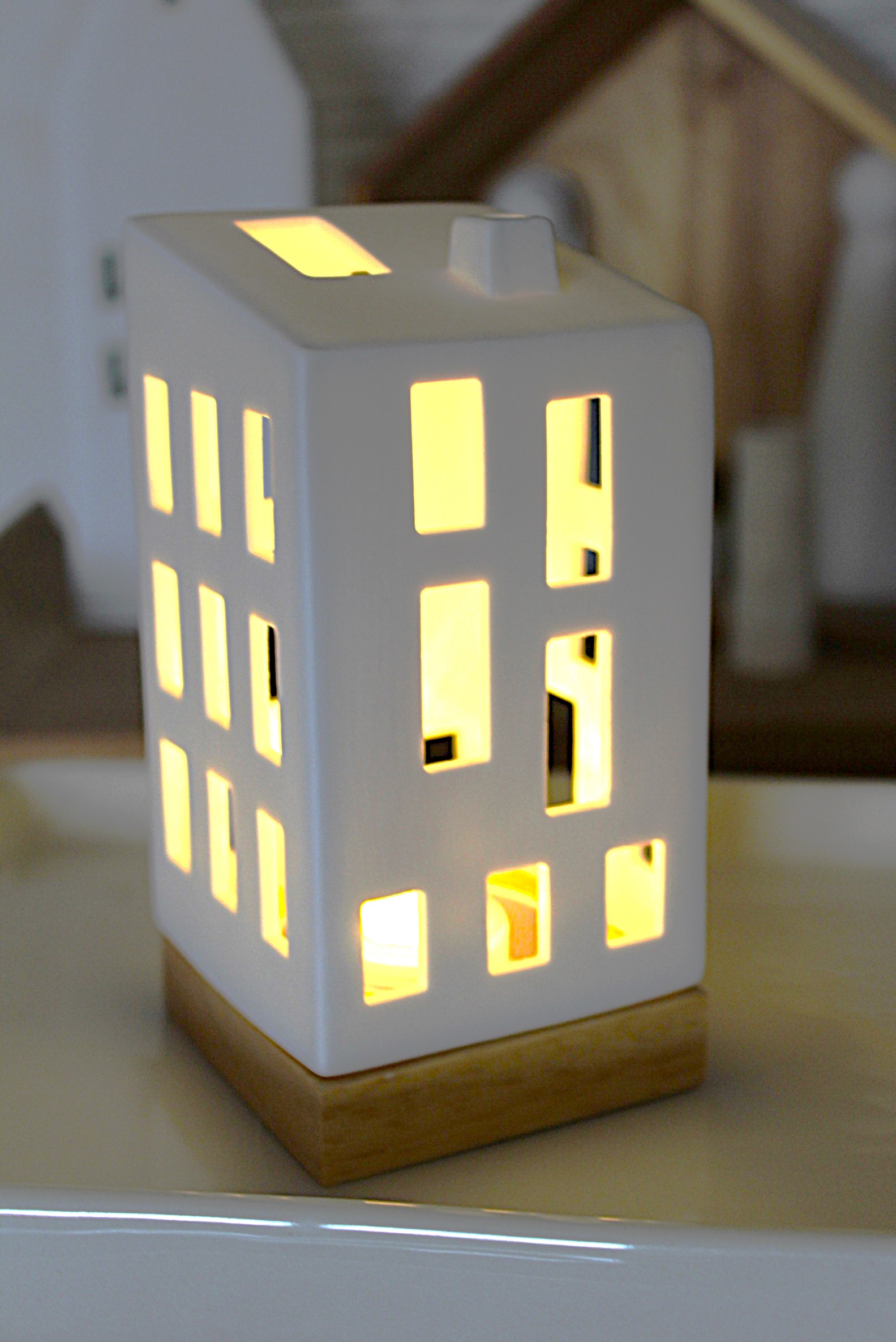 Moderner hausförmiger Kerzenhalter aus weißer Keramik mit beleuchteten Fenstern, der auf einem Holzsockel ruht und ein warmes, einladendes Licht erzeugt, das sich perfekt für die Inneneinrichtung und das Ambiente eignet.