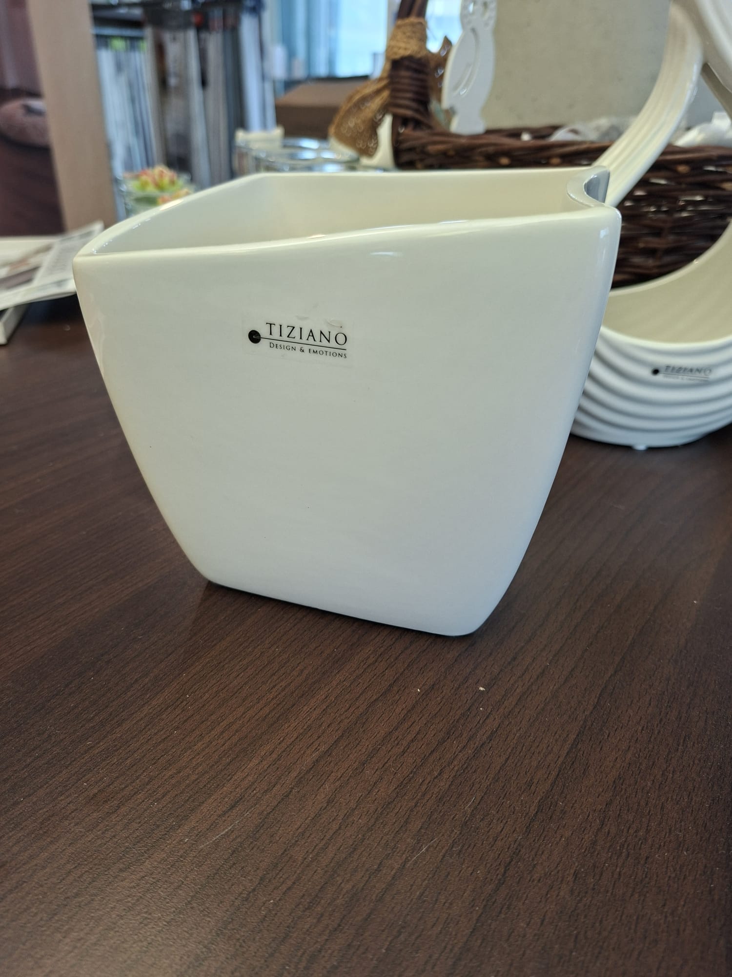 Ein weißer, quadratischer Keramikpflanzer von Tiziano Design & Emotions steht auf einem dunklen Holztisch, im Hintergrund sind ein verschwommener Weidenkorb und andere Einrichtungsgegenstände zu sehen.