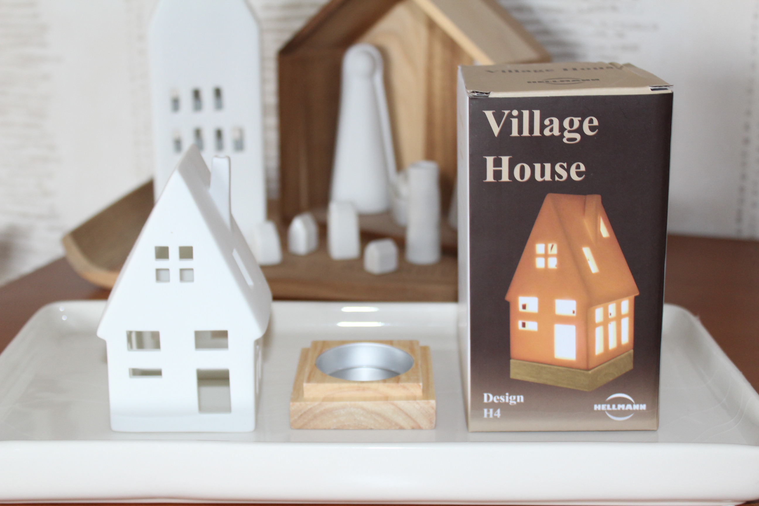 Ein weißes Dorfhaus aus Keramik, ein Teelichthalter aus Holz und eine „Village House“-Produktbox werden auf einem weißen Tablett präsentiert, mit anderen dekorativen Häusern im Hintergrund.