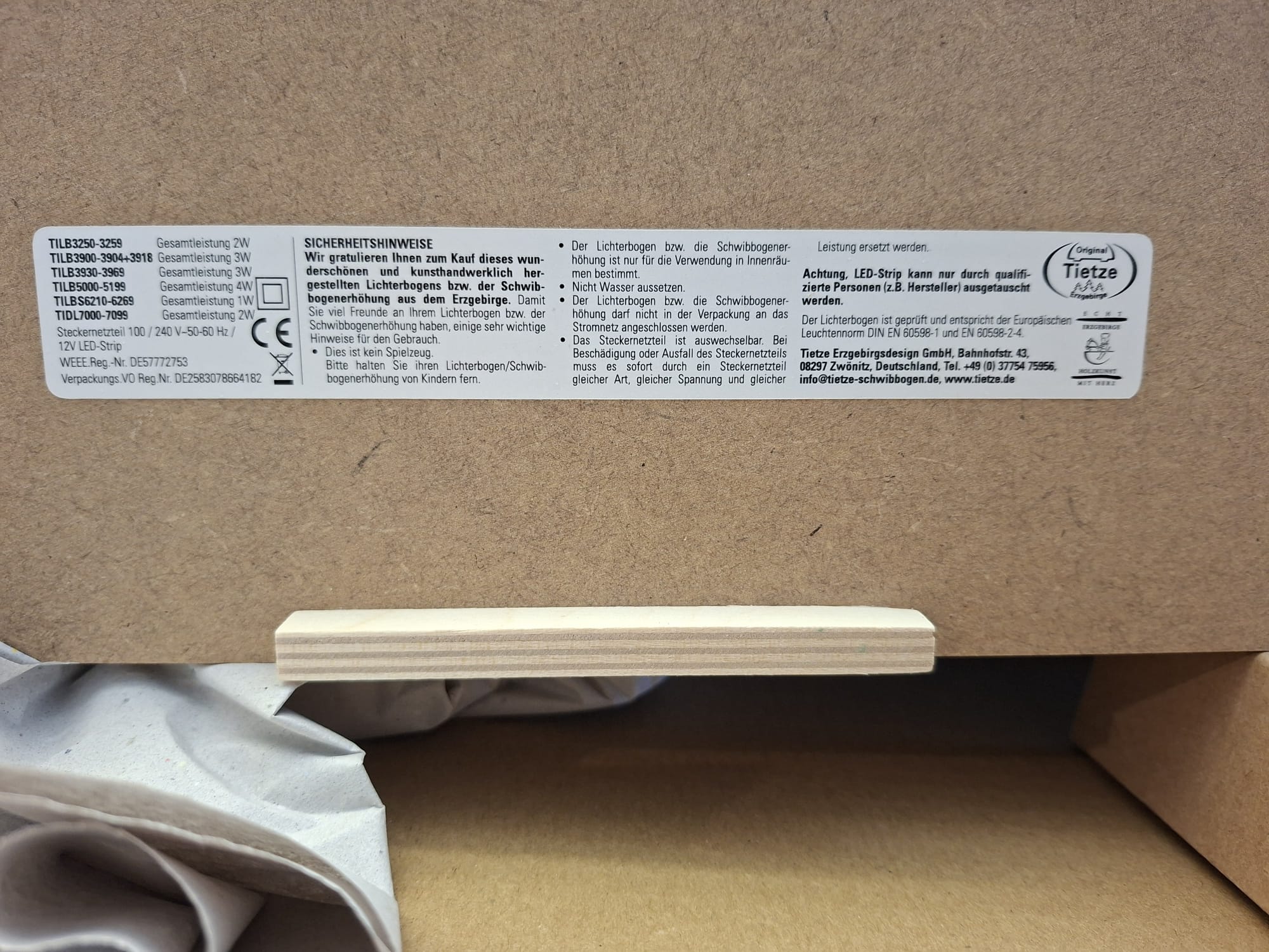 Eine Nahaufnahme eines Produktinformationsetiketts auf einer Holzoberfläche mit technischen Spezifikationen, Sicherheitshinweisen auf Deutsch und dem Firmenlogo von Tietze Erzgebirgsdesign, darunter ist ein kleines hölzernes Stützstück sichtbar.