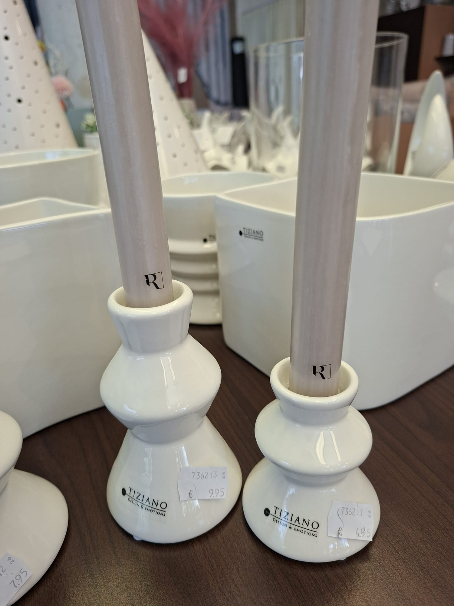 Zwei weiße Tiziano-Kerzenhalter aus Keramik mit hohen, hellen Spitzkerzen auf einer Holzoberfläche, umgeben von anderen weißen Keramik-Wohnaccessoires in einer Verkaufspräsentation.