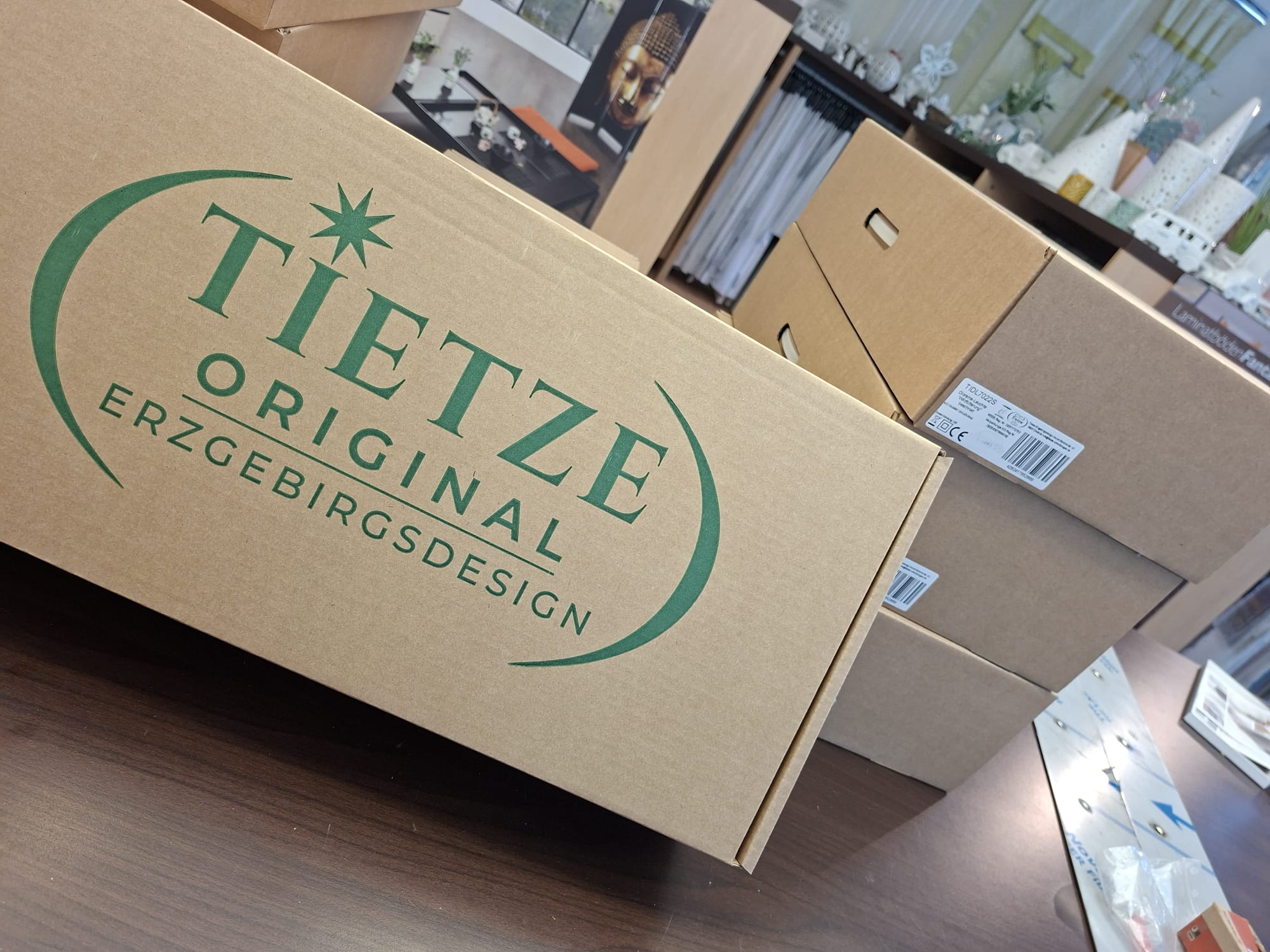 Ein Stapel brauner Versandkartons aus Pappe mit dem grünen Logo von Tietze Original Erzgebirgsdesign, platziert auf einem dunklen Holztisch in einer Einzelhandels- oder Lagerumgebung.