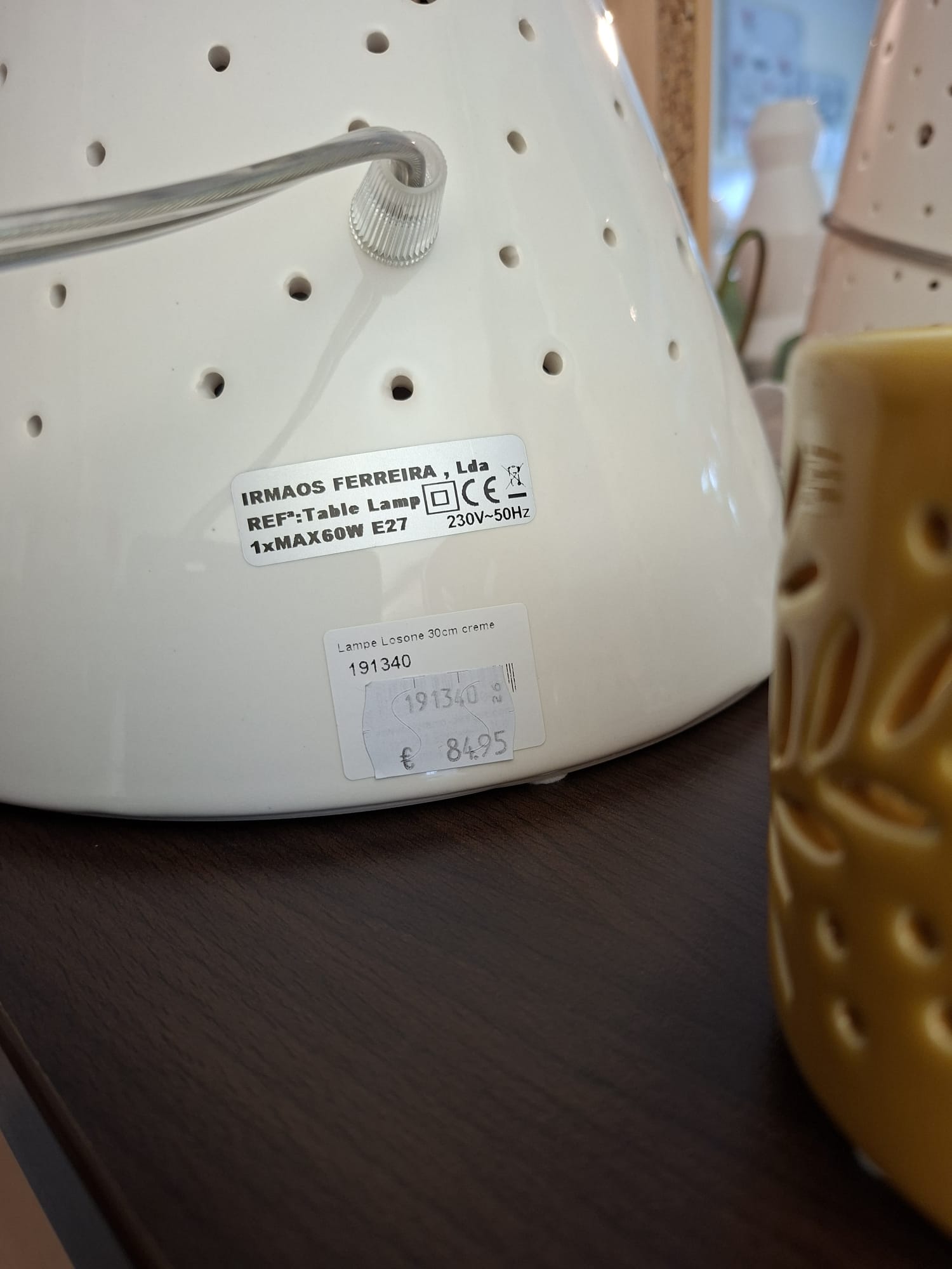 Eine Nahaufnahme eines weißen, perforierten Tischlampensockels mit einem Produktetikett mit technischen Spezifikationen und einem Preisschild von 84,95 Euro, der auf einer dunklen Holzoberfläche neben einem gelben Dekorationsobjekt ruht.