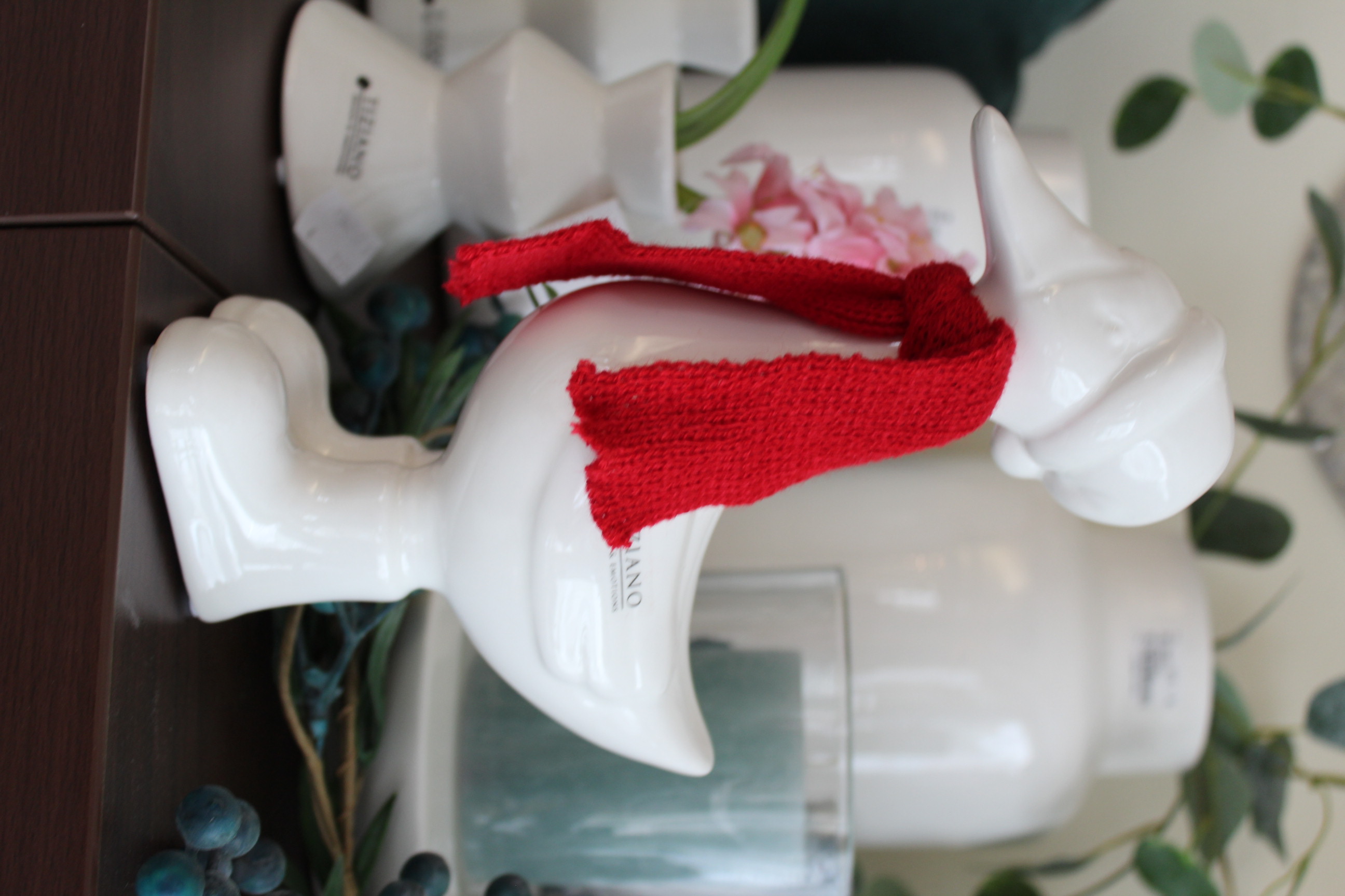 Eine weiße Keramik-Entenfigur mit rotem Strickschal und passender weißer Mütze, ausgestellt auf einer dunklen Oberfläche mit dekorativen Vasen, Grünpflanzen und einer Kerze im Hintergrund.