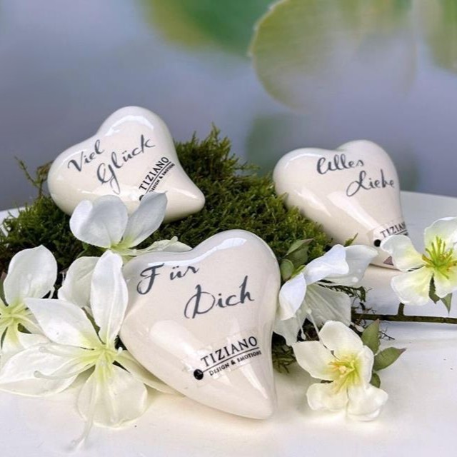 Drei cremefarbene Keramikherzen mit den Aufschriften 'Viel Glück', 'Für Dich' und 'Alles Liebe' liegen auf Moos zwischen weißen Blüten.