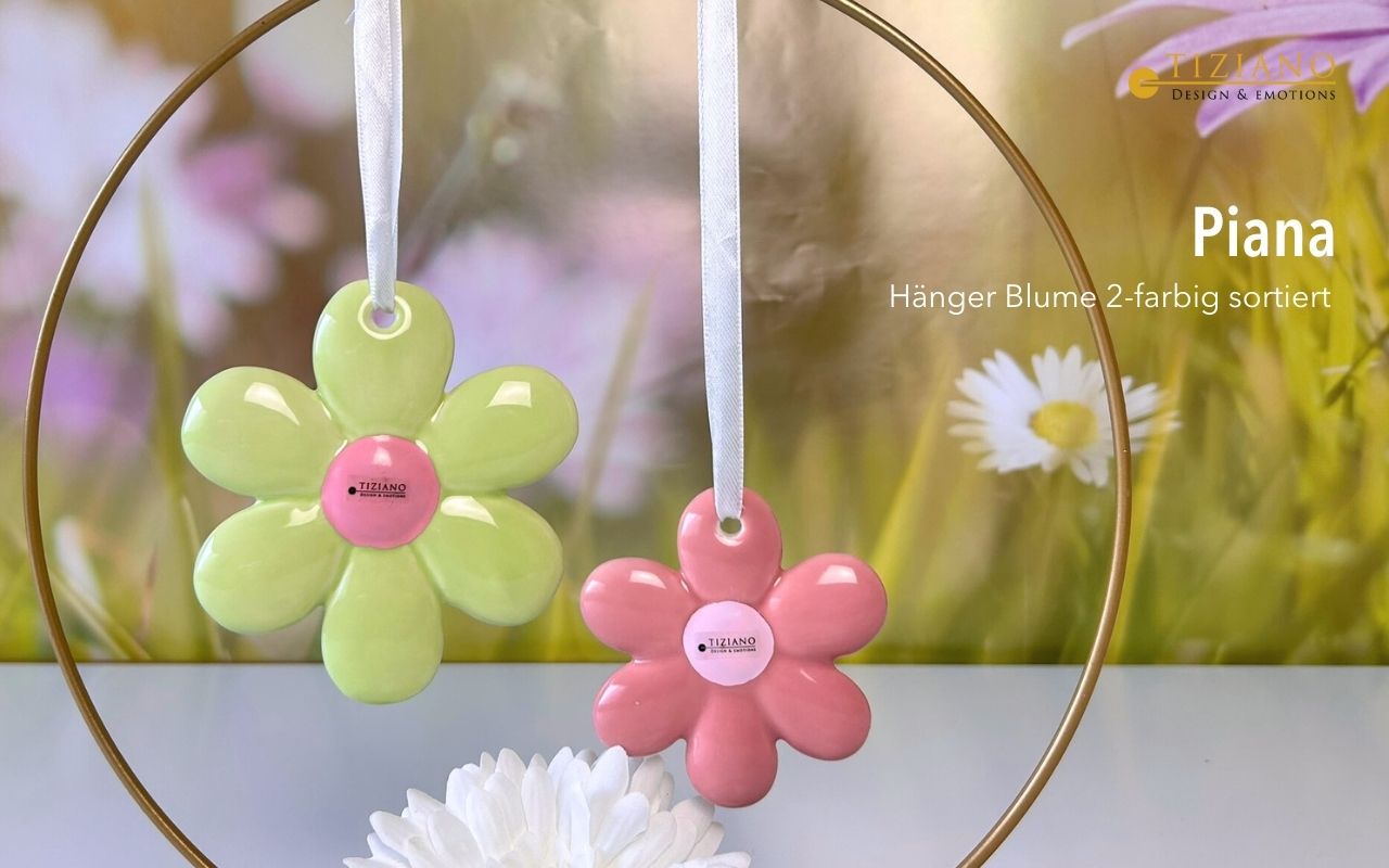 Zwei farbenfrohe, blütenförmige Ornamente in Grün und Rosa, die an einem weißen Band vor einem verschwommenen Blumenhintergrund hängen und ein dekoratives Design von Tiziano zeigen.