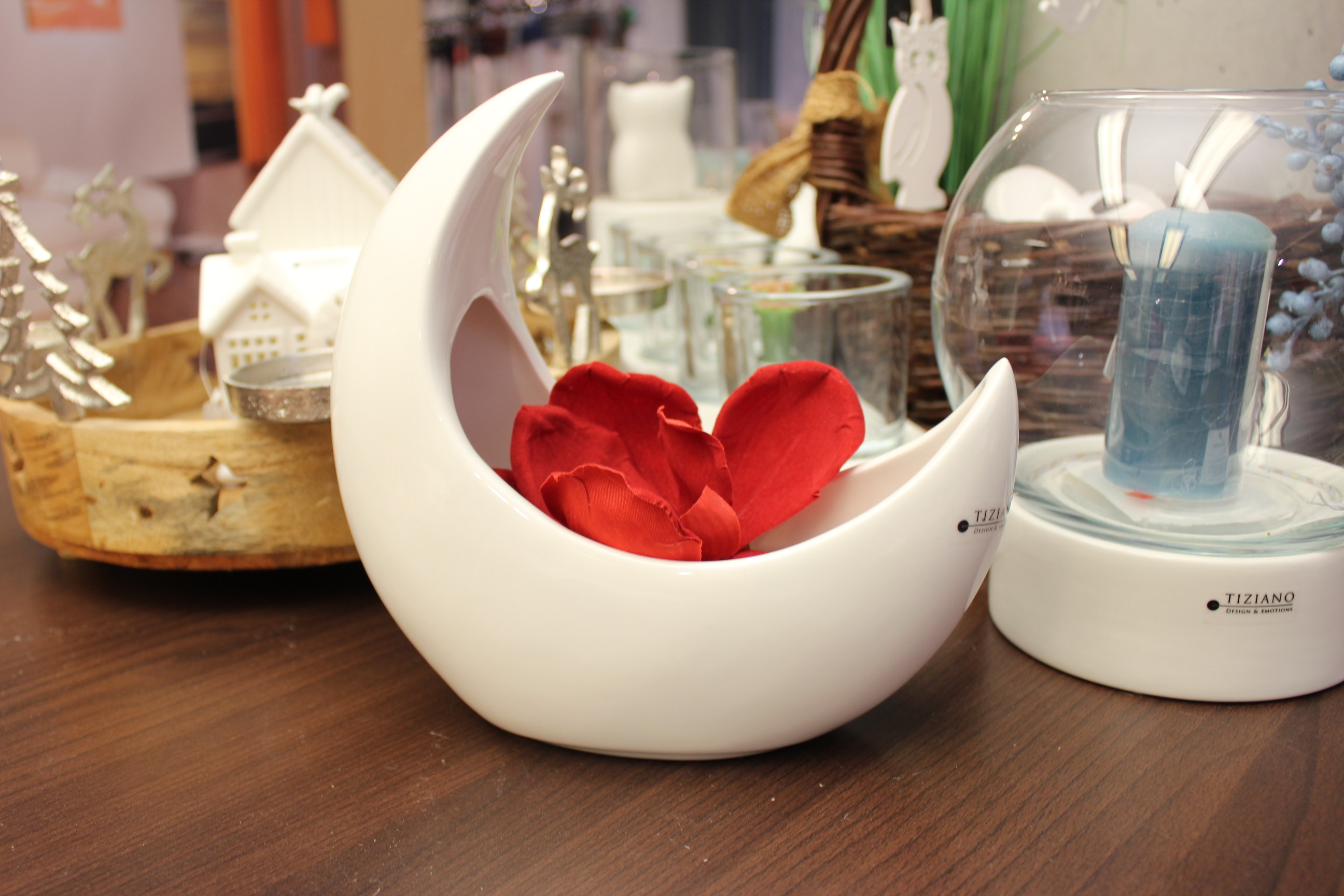 Ein halbmondförmiger cremefarbener Keramik-Übertopf von Tiziano Design & Emotions Marina mit einer leuchtend roten Blume, ausgestellt auf einer Holzoberfläche mit anderen dekorativen Feiertagsgegenständen im Hintergrund.